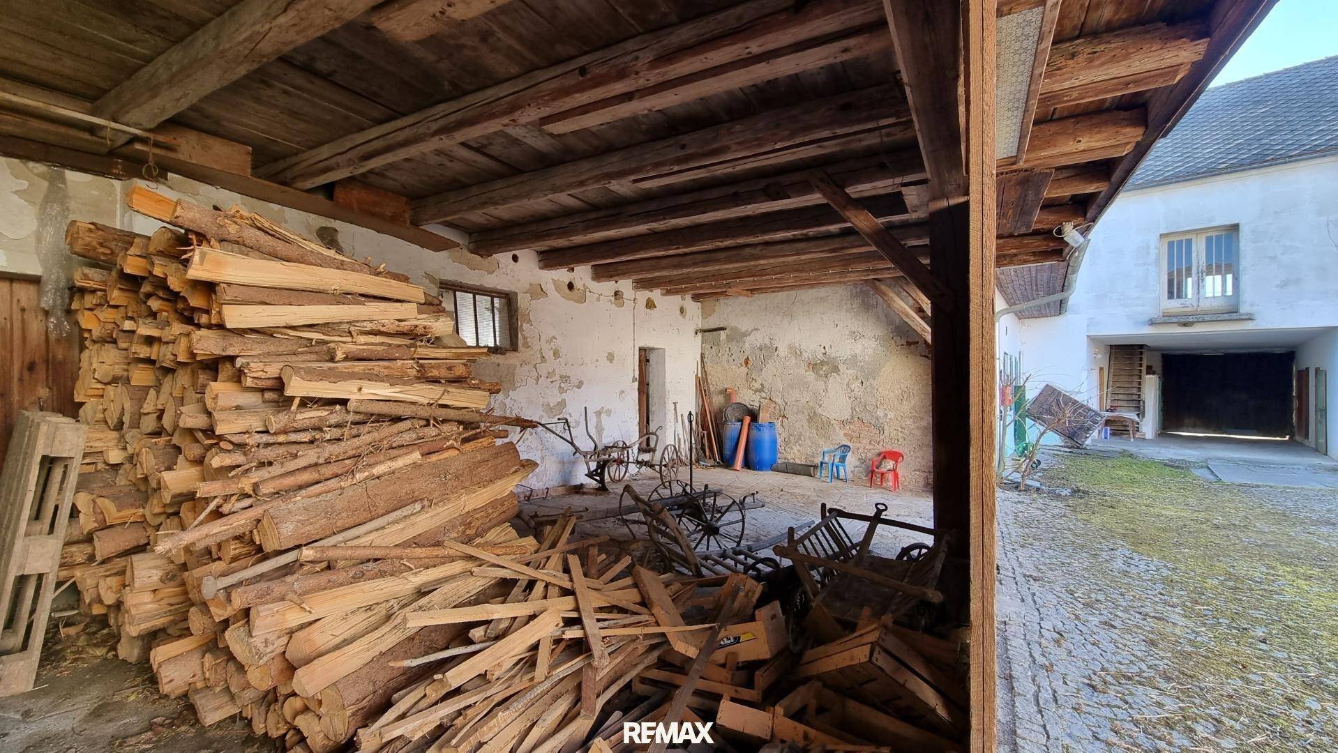 Geräumige Scheune mit großen Holzbalken, gestapeltem Holz und Blick auf den Innenhof.