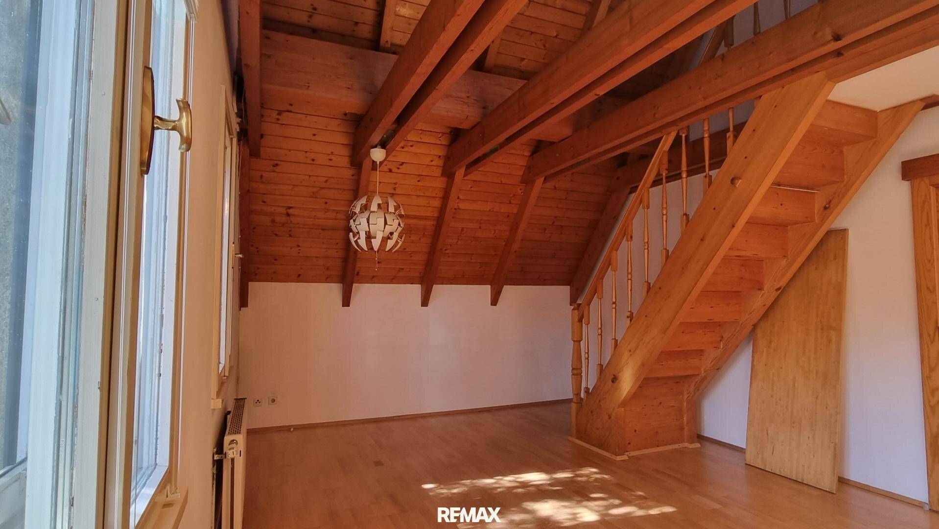 Geräumiger Wohnbereich im Dachgeschoss mit Holzbalken, Treppe und großen Fenstern.