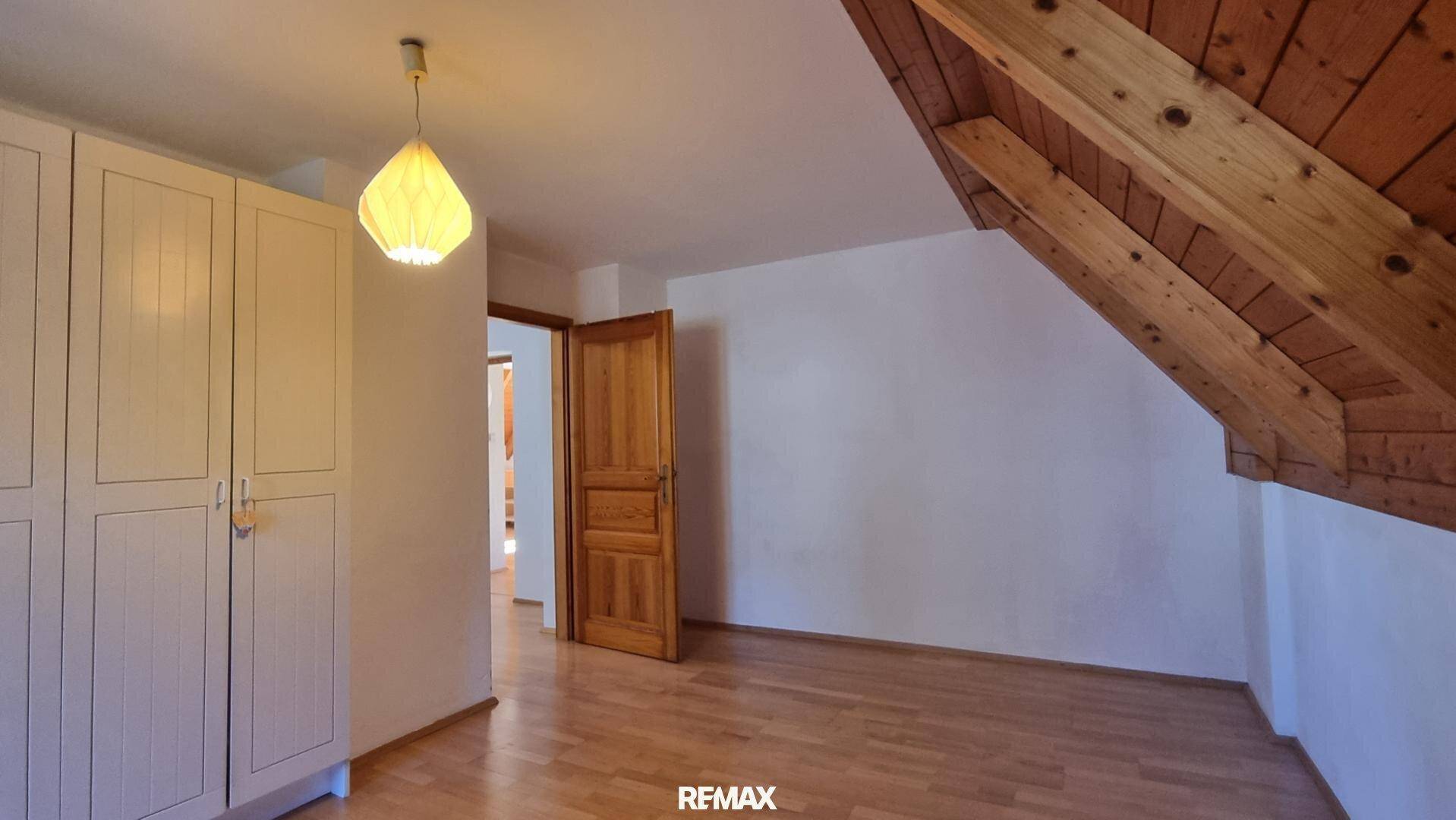 Dachzimmer mit Holzboden, Dachschräge und weißem Kleiderschrank, mit Zugang zu weiteren Räumen.