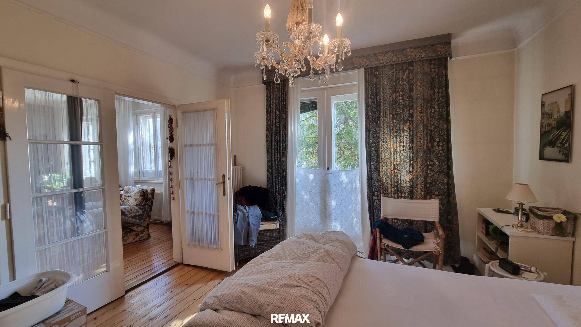 Schlafzimmer mit Holzboden, Kronleuchter und Zugang zu einem weiteren Raum und Balkon.