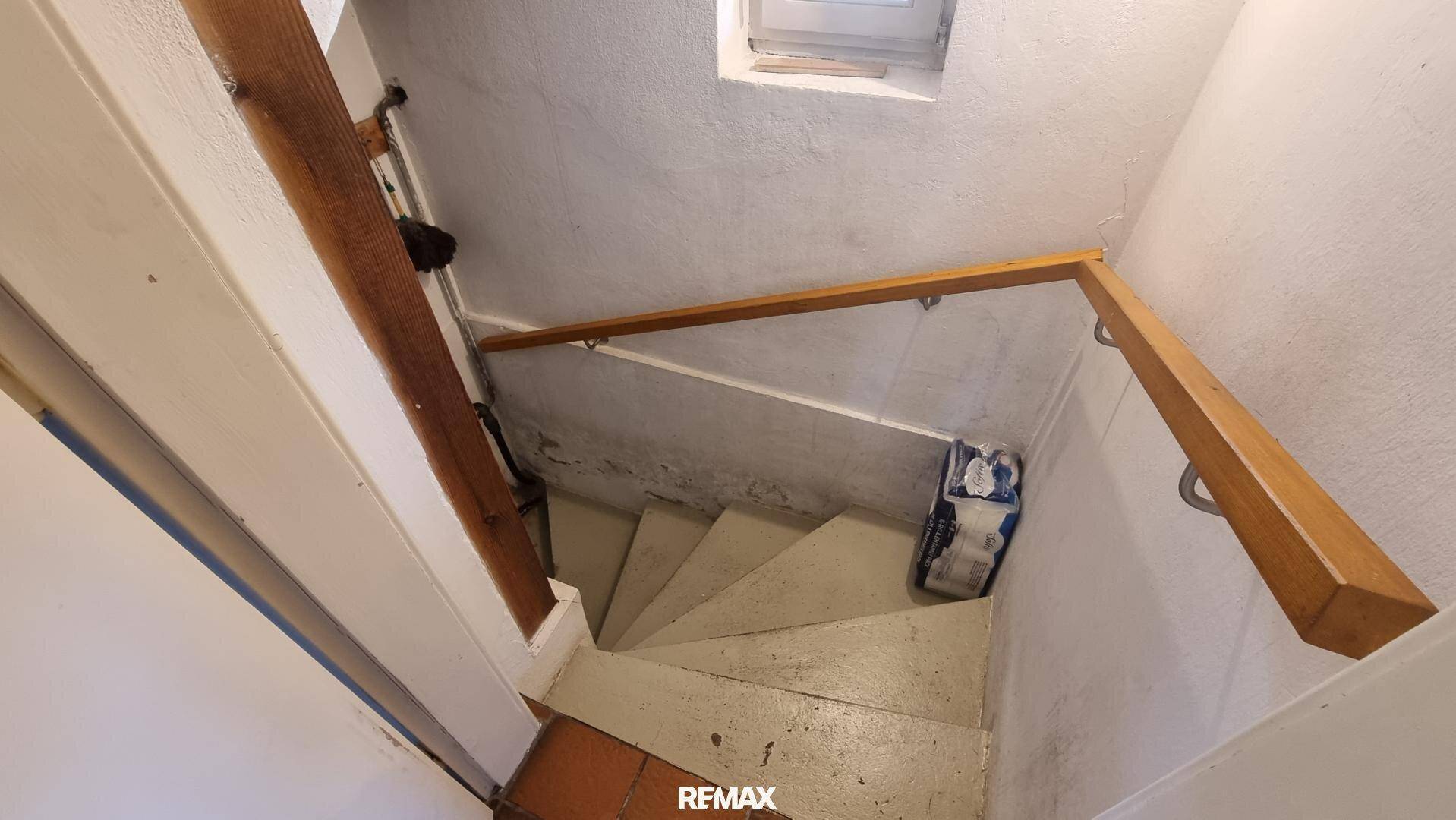 Schmale Treppe mit Holzgeländer und weiß gestrichenen Wänden, die in ein Obergeschoss führt.