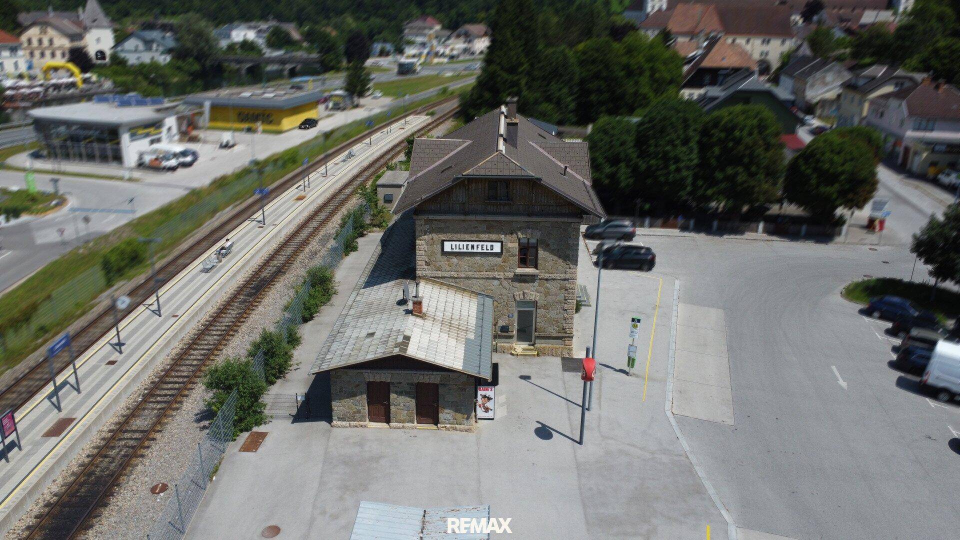 Luftaufnahme des Bahnhofs Lilienfeld mit Bahngleisen, Parkplätzen und umliegender Bebauung vor grüner Bergkulisse.