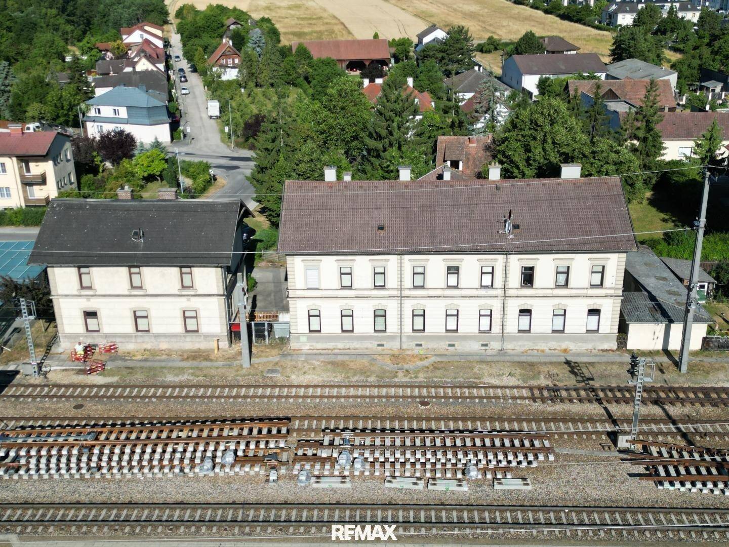 Weitwinkelaufnahme von zwei Gebäuden entlang der Bahngleise, umgeben von ländlicher Bebauung und Feldern.