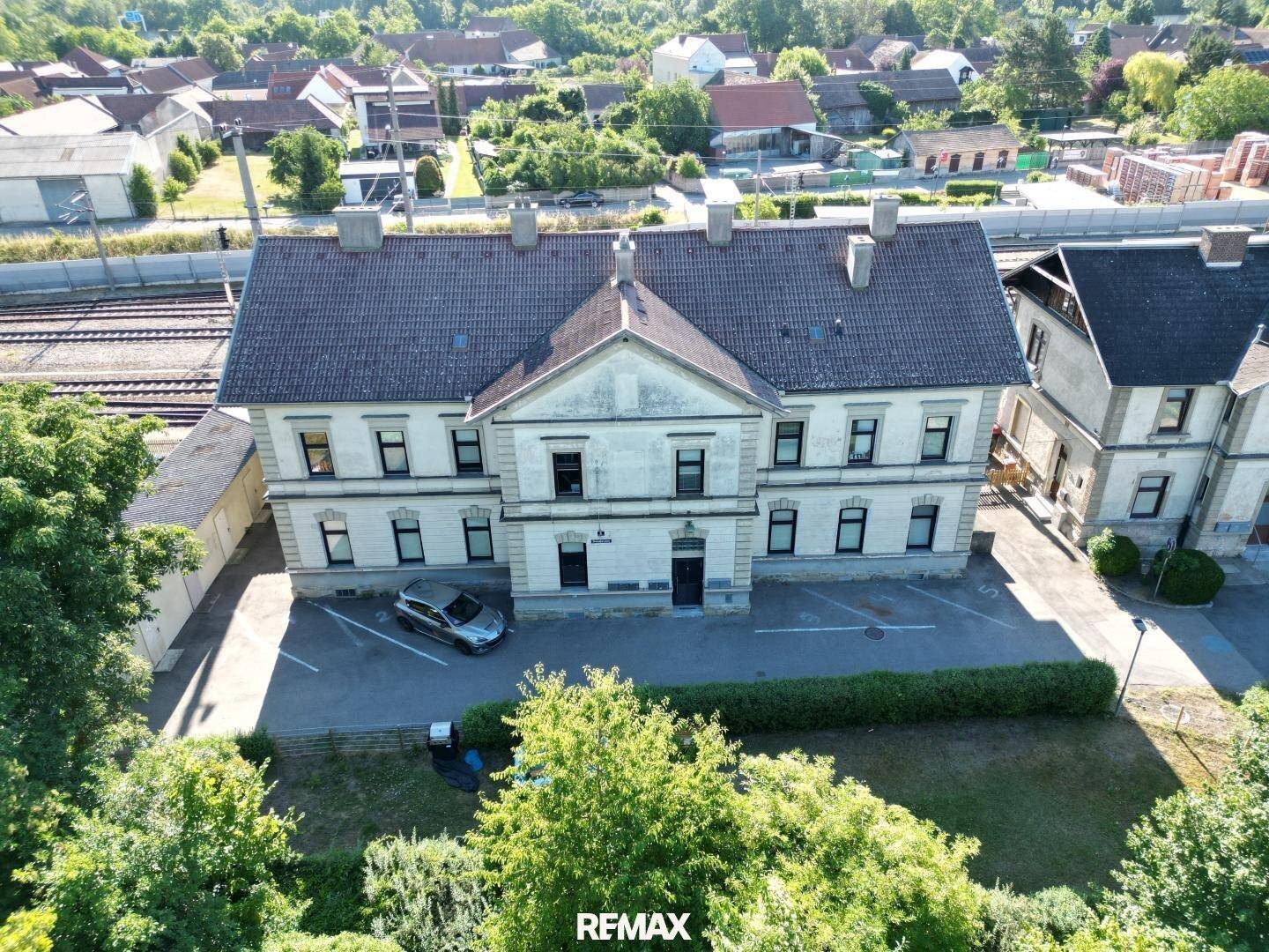 Draufsicht auf zwei ältere Gebäude mit Satteldächern, umgeben von Bäumen und einem Parkplatz.