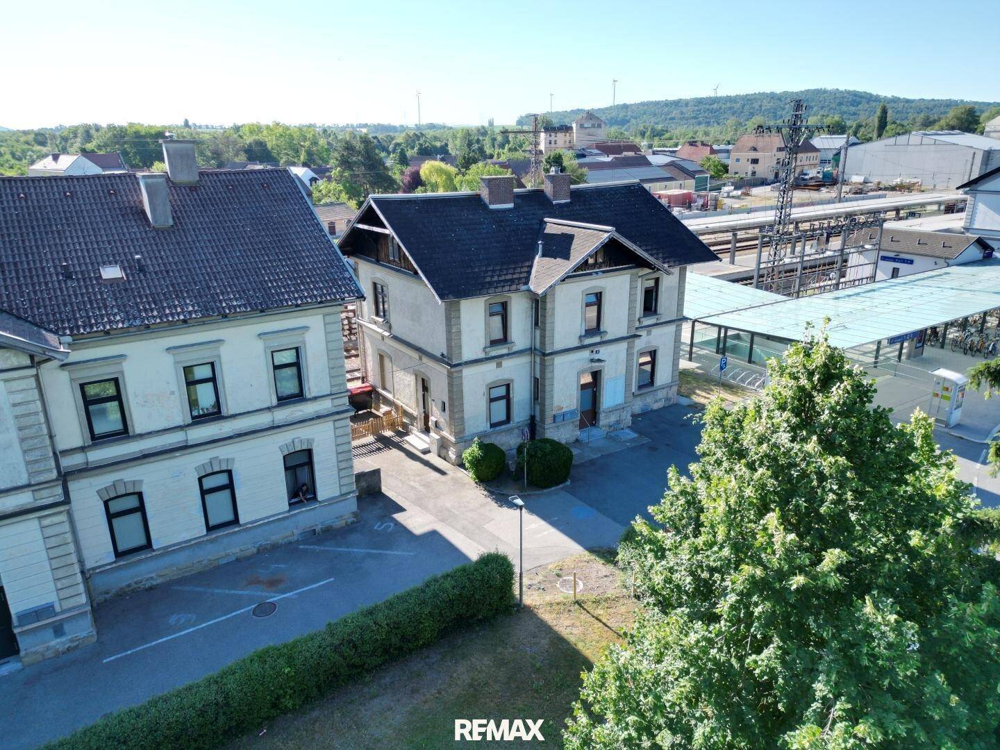 Blick von oben auf zwei Gebäude neben Bahngleisen, mit grüner Landschaft im Hintergrund und klarem Himmel.