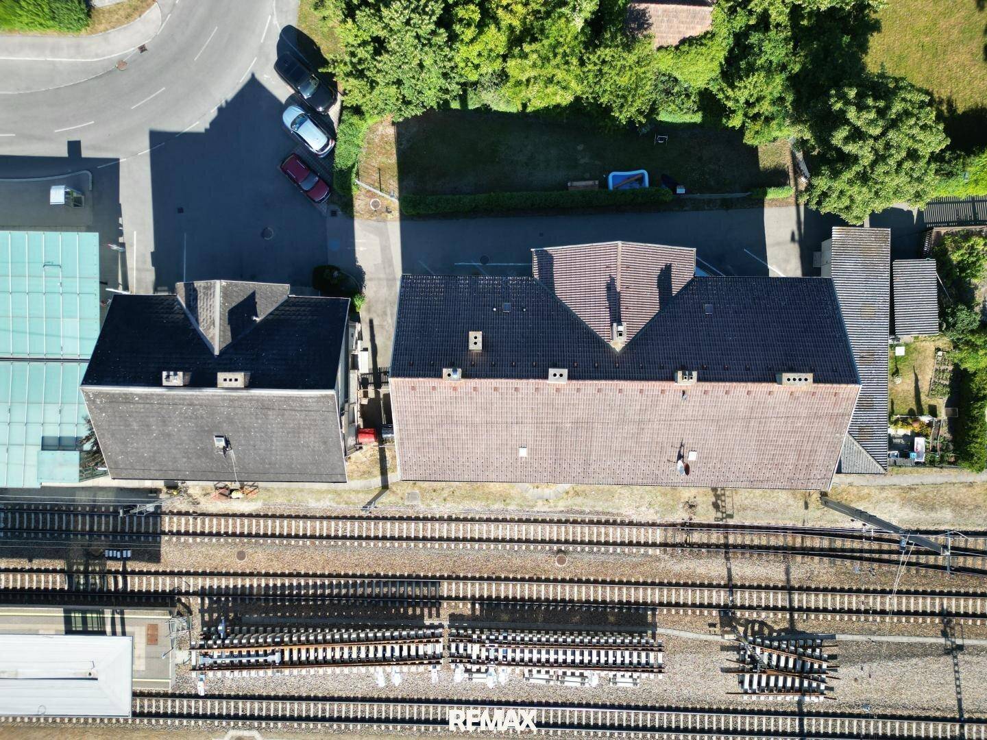 Senkrechte Luftaufnahme der Dächer zweier Gebäude, angrenzend an Bahngleise und eine Straße.