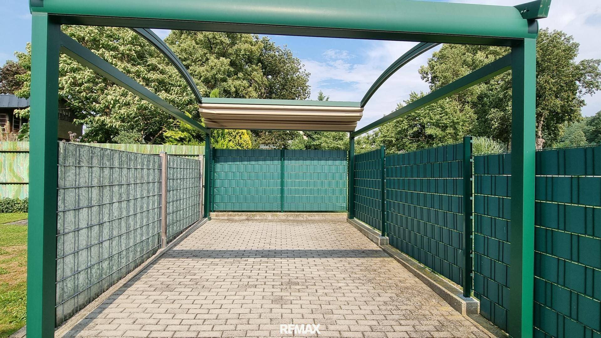 Überdachter Carport mit gepflastertem Boden und grünen Sichtschutzelementen.