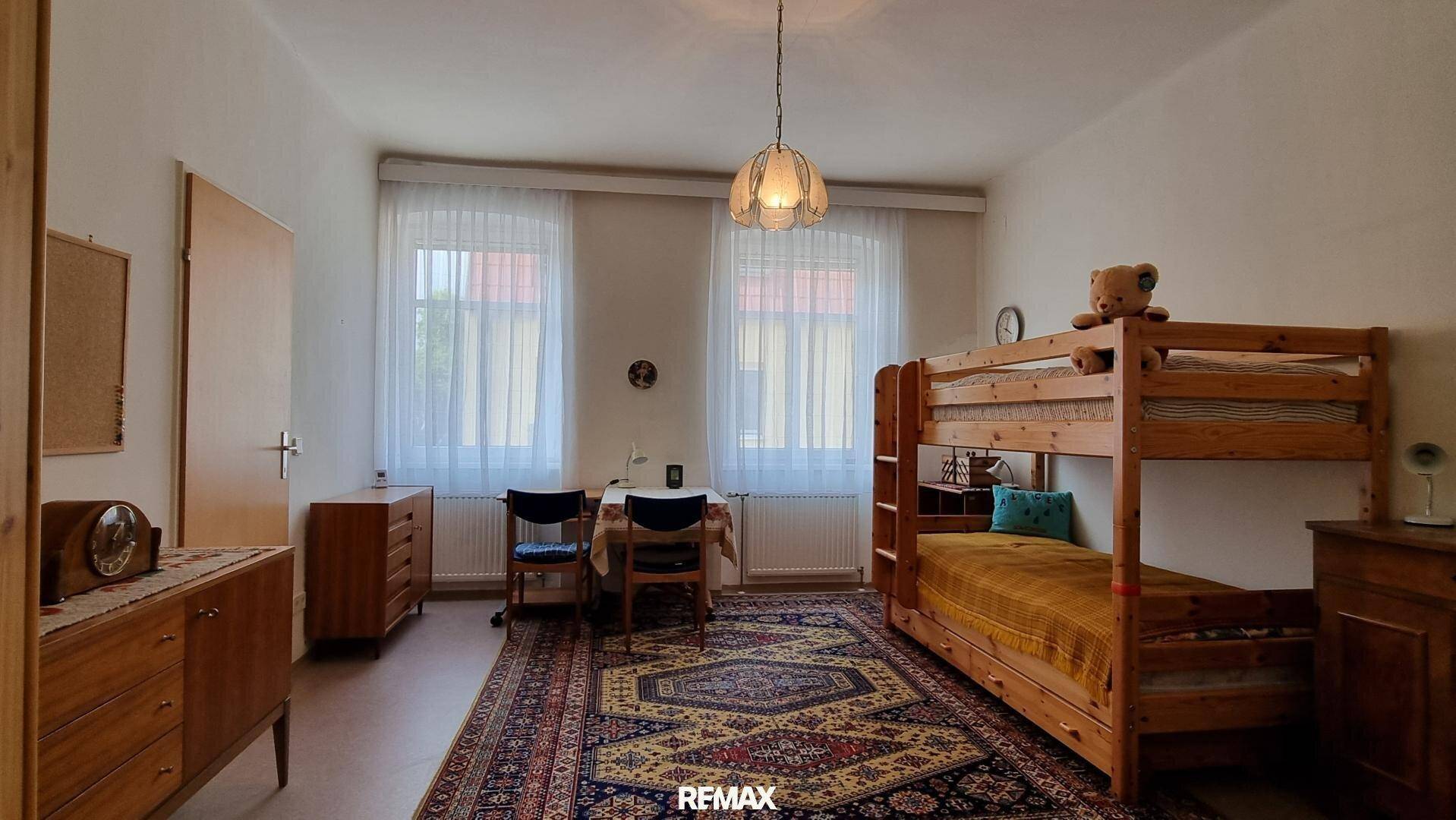 Kinderzimmer mit Etagenbett, Holzmöbeln und Teppichboden, hell durch zwei Fenster.
