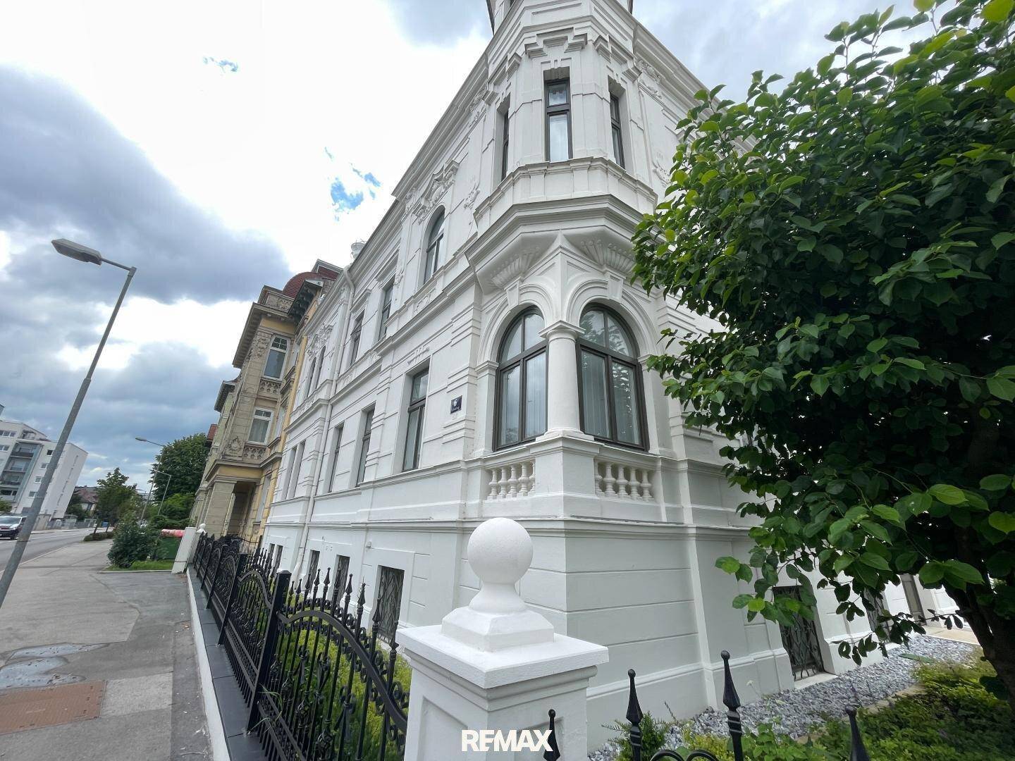 Eckansicht der weißen, verzierten Gebäudefassade mit großen Fenstern und einem Baum.