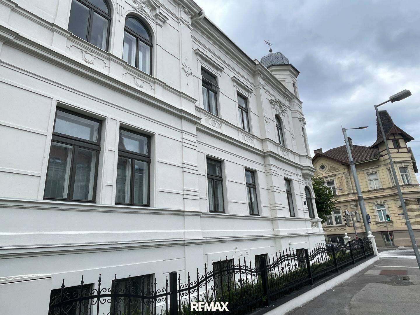 Historische weiße Fassade eines Gebäudes mit verzierten Details und dunklen Fenstern.