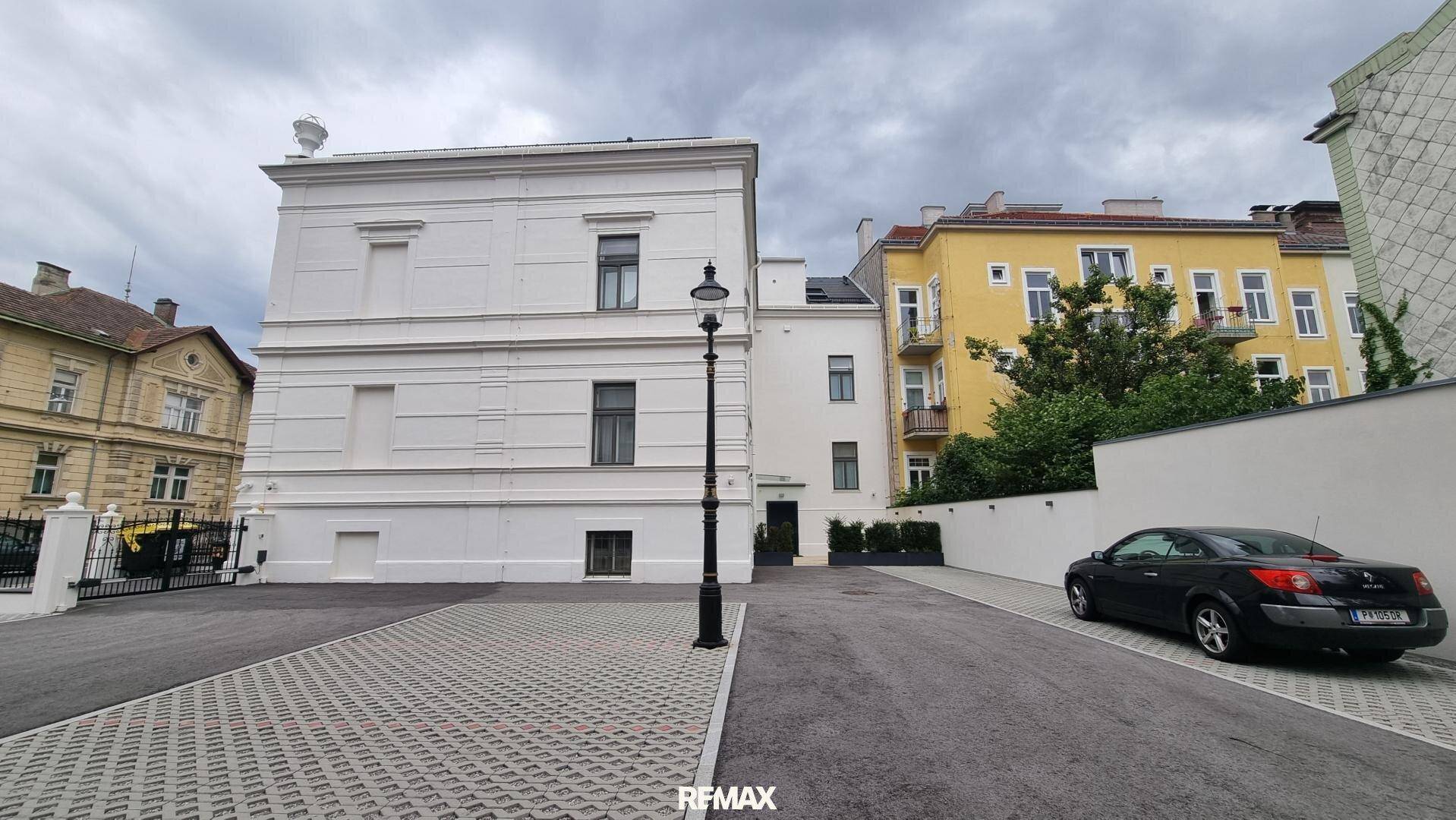 Rückansicht eines weißen Gebäudes mit Parkplatz und einem weiteren historischen Gebäude im Hintergrund.