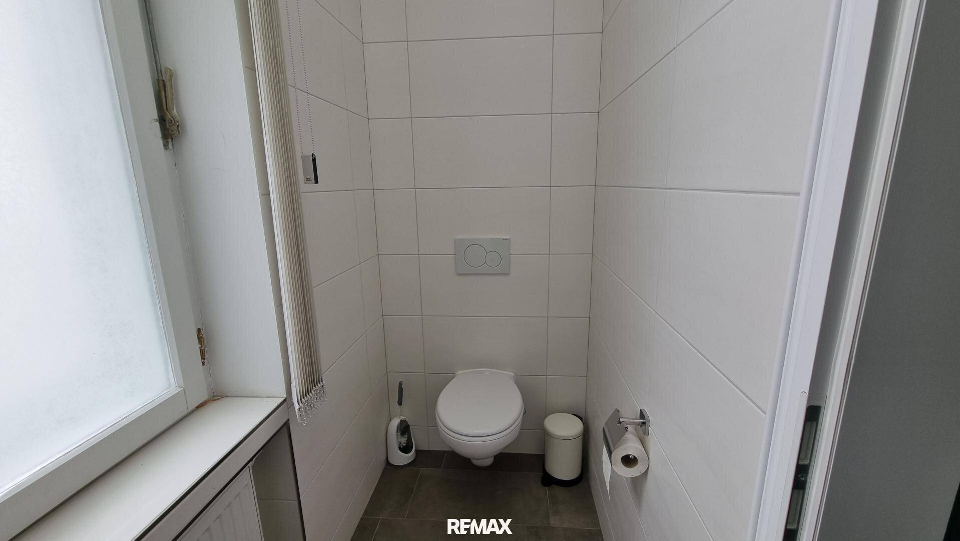 Helles WC mit weißen Wandfliesen, einer Toilette und einem Fenster für natürliche Belüftung.
