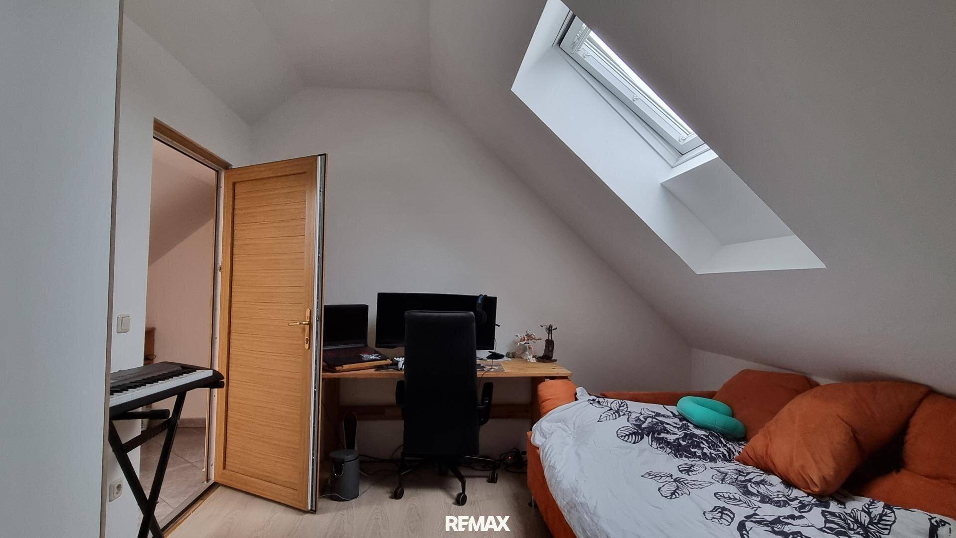 Dachgeschoss-Büro mit Schreibtisch, Computer und Sofa, beleuchtet durch ein Dachfenster und mit schrägen Wänden.