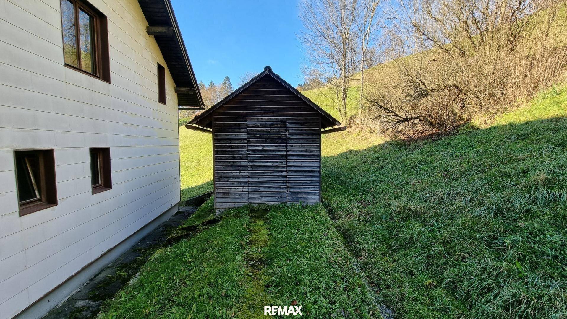 Seitenansicht eines Hauses mit weißer Verkleidung und einem kleinen Holzschuppen auf einer grünen Wiese.