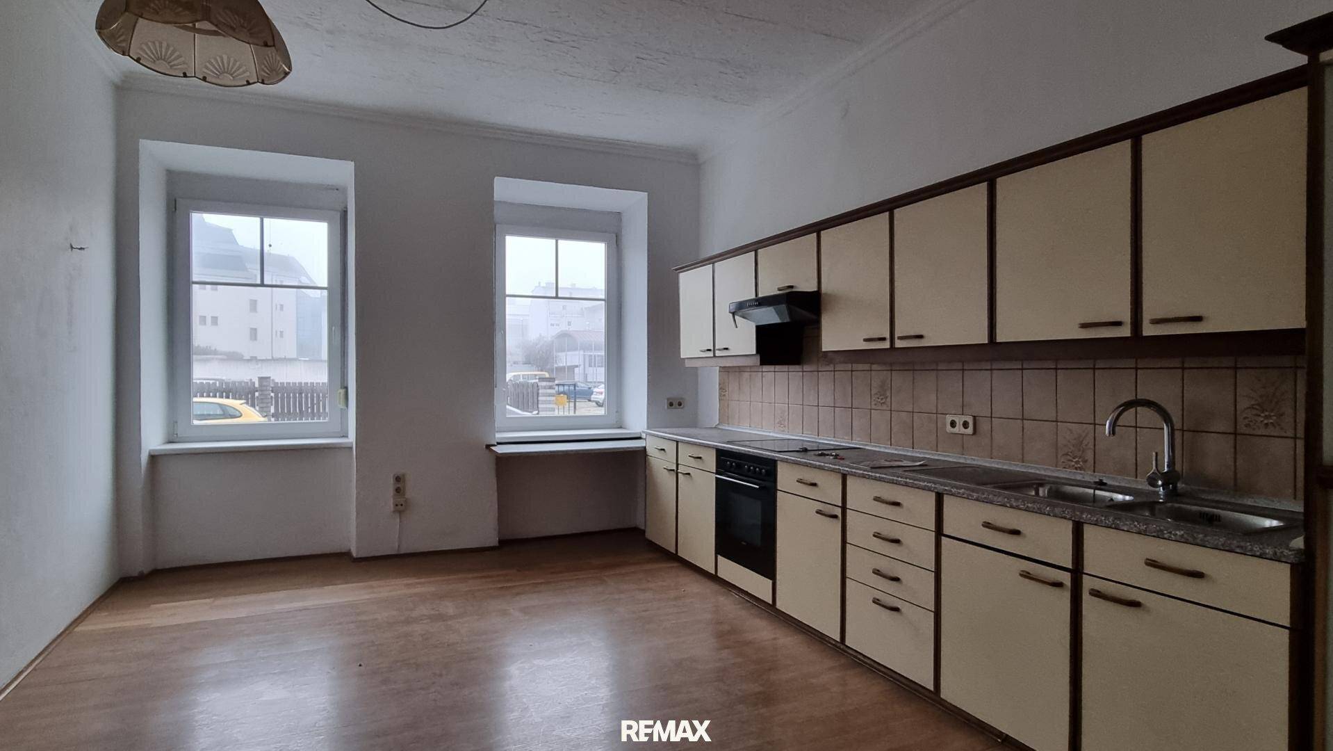 Küche mit älteren Einbauschränken, Spüle und Blick auf die Straße durch zwei Fenster.
