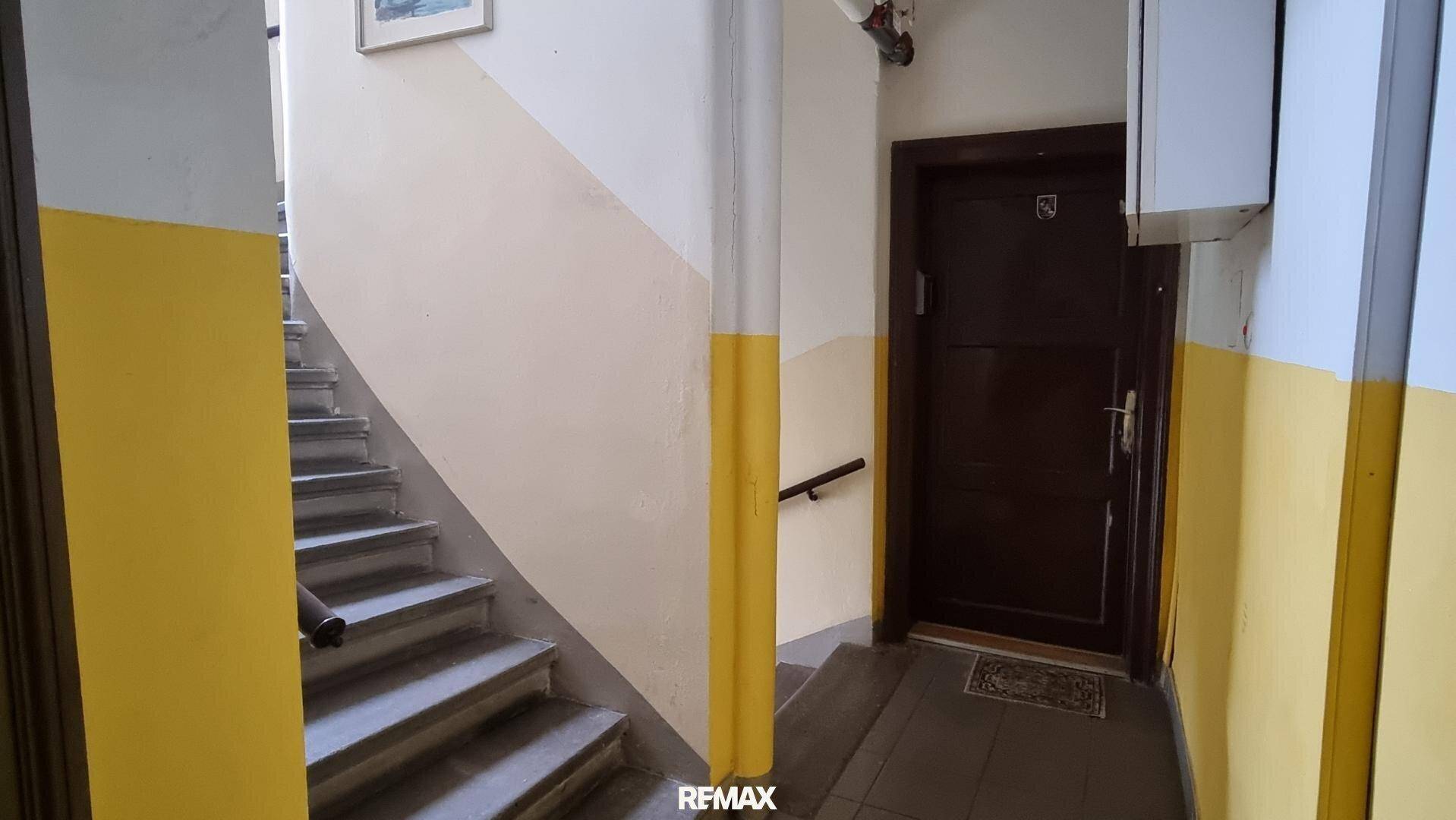 Treppenhaus mit gelb-weißen Wänden, Fliesenboden und einer dunklen Eingangstür zu einer Wohneinheit.