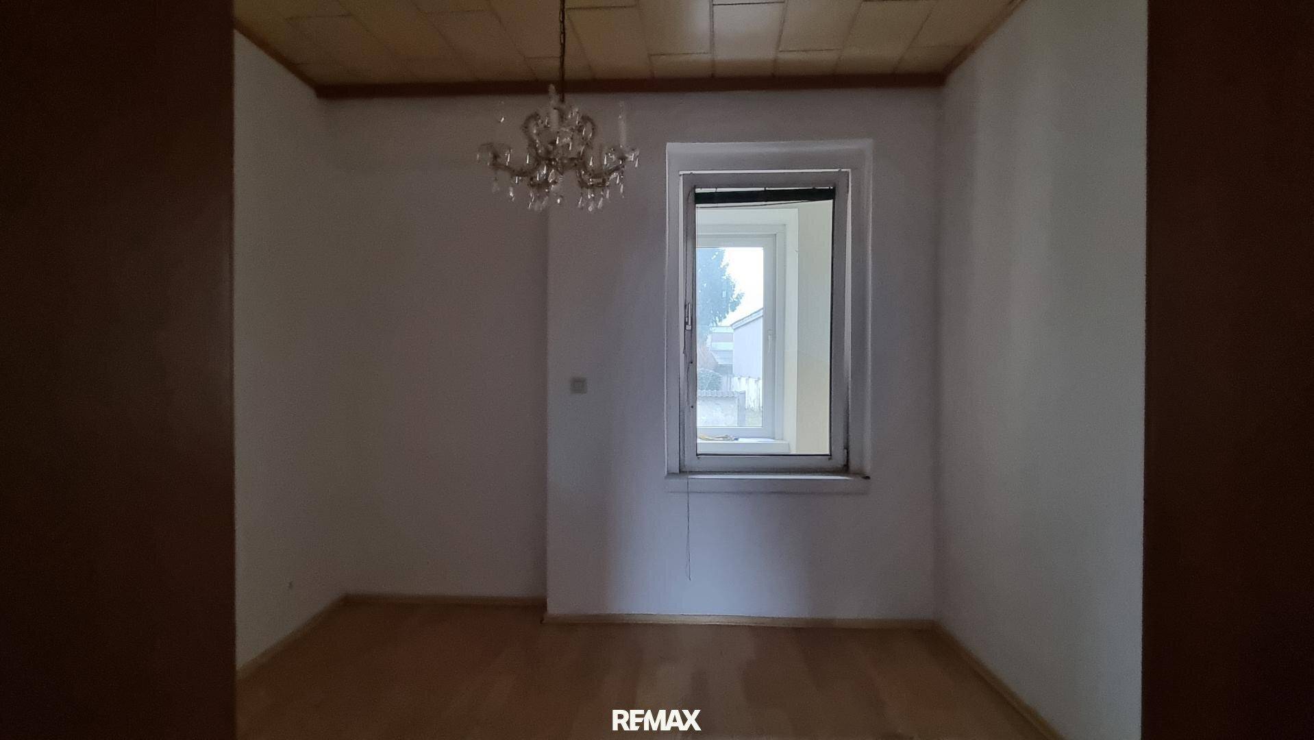 Helles Zimmer mit Holzboden, Kronleuchter und einem großen Fenster, das viel Licht hereinlässt.