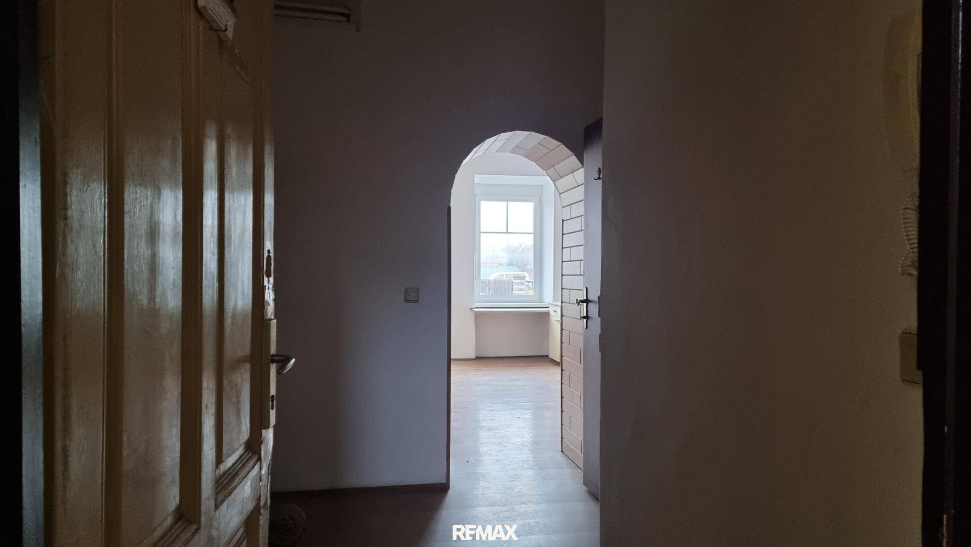 Flur mit Holztür und einem Bogen, der zu einem hellen Raum mit Fenster führt und einladend wirkt.