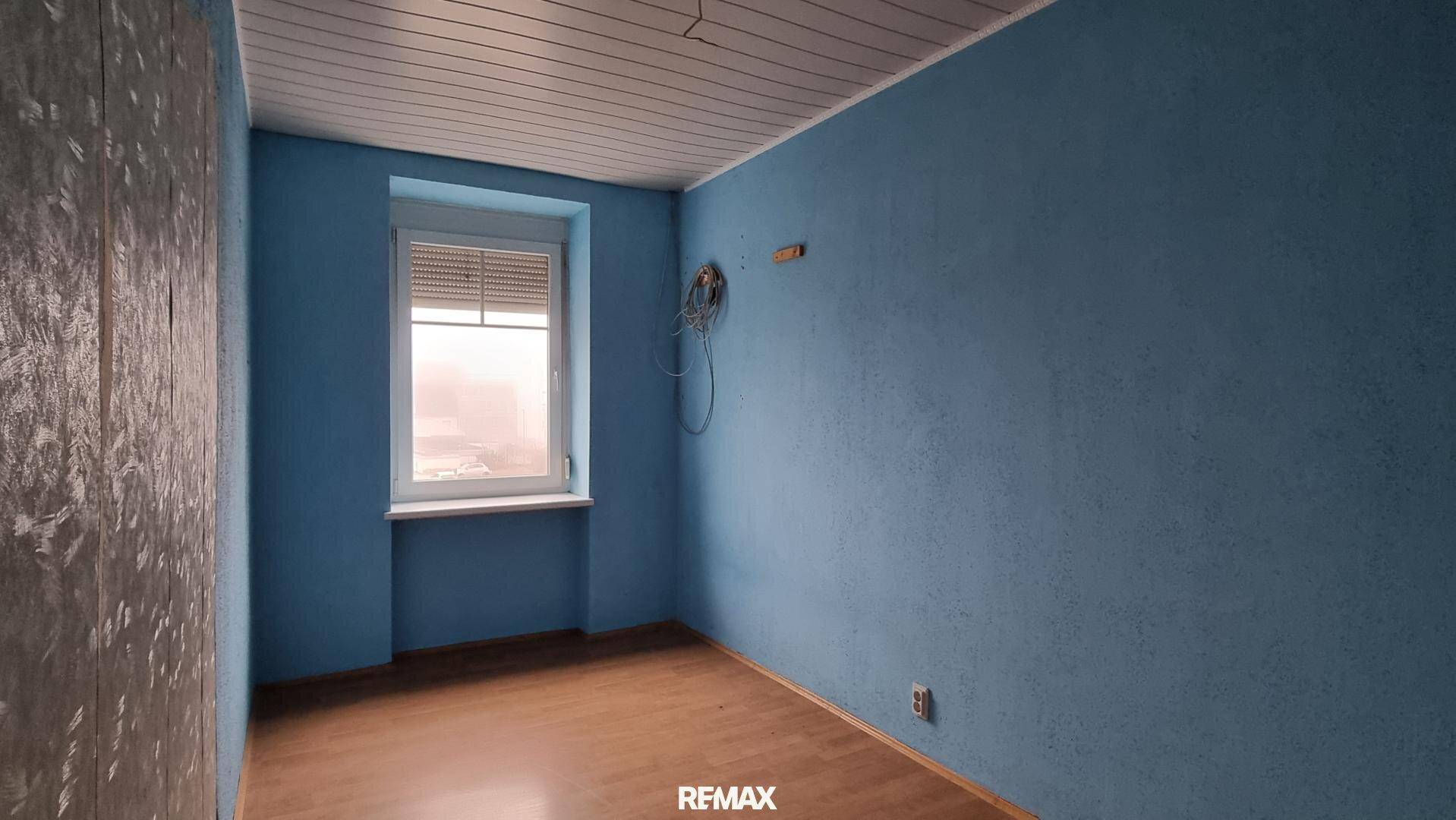 Kleines Zimmer mit blauen Wänden, einem Fenster und Holzdeckenverkleidung, bereit zur individuellen Gestaltung.