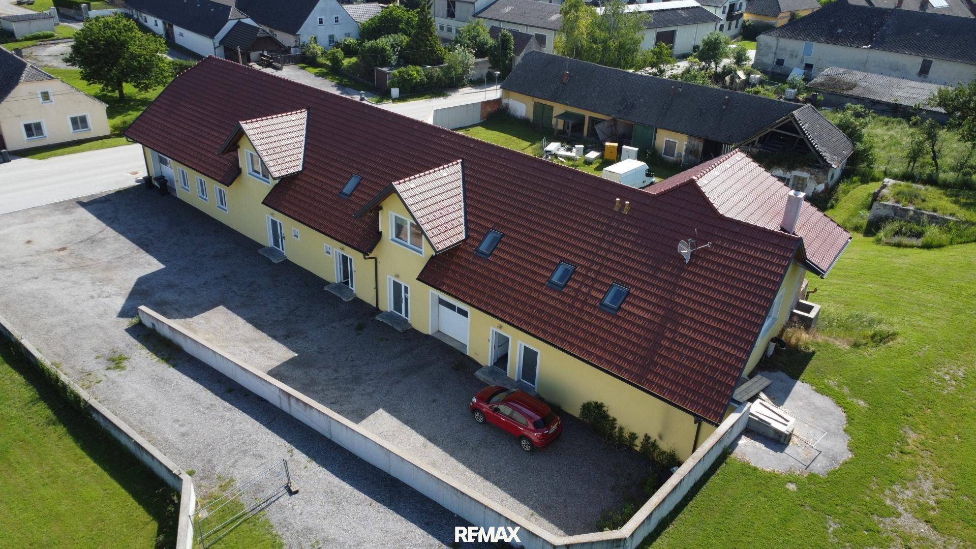 Luftaufnahme eines langen, gelben Gebäudes mit rotem Ziegeldach und mehreren Dachgauben in ländlicher Umgebung.