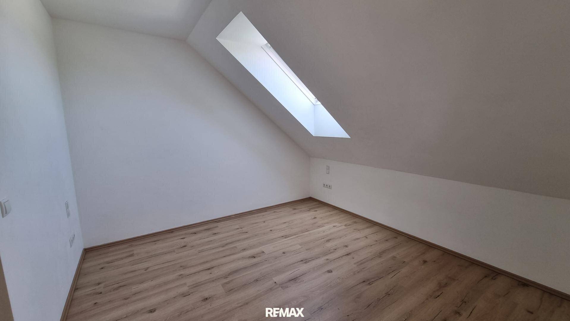 Schlafzimmer mit hellem Laminatboden, weißen Wänden und einem einzelnen Dachfenster, das den Raum erhellt.