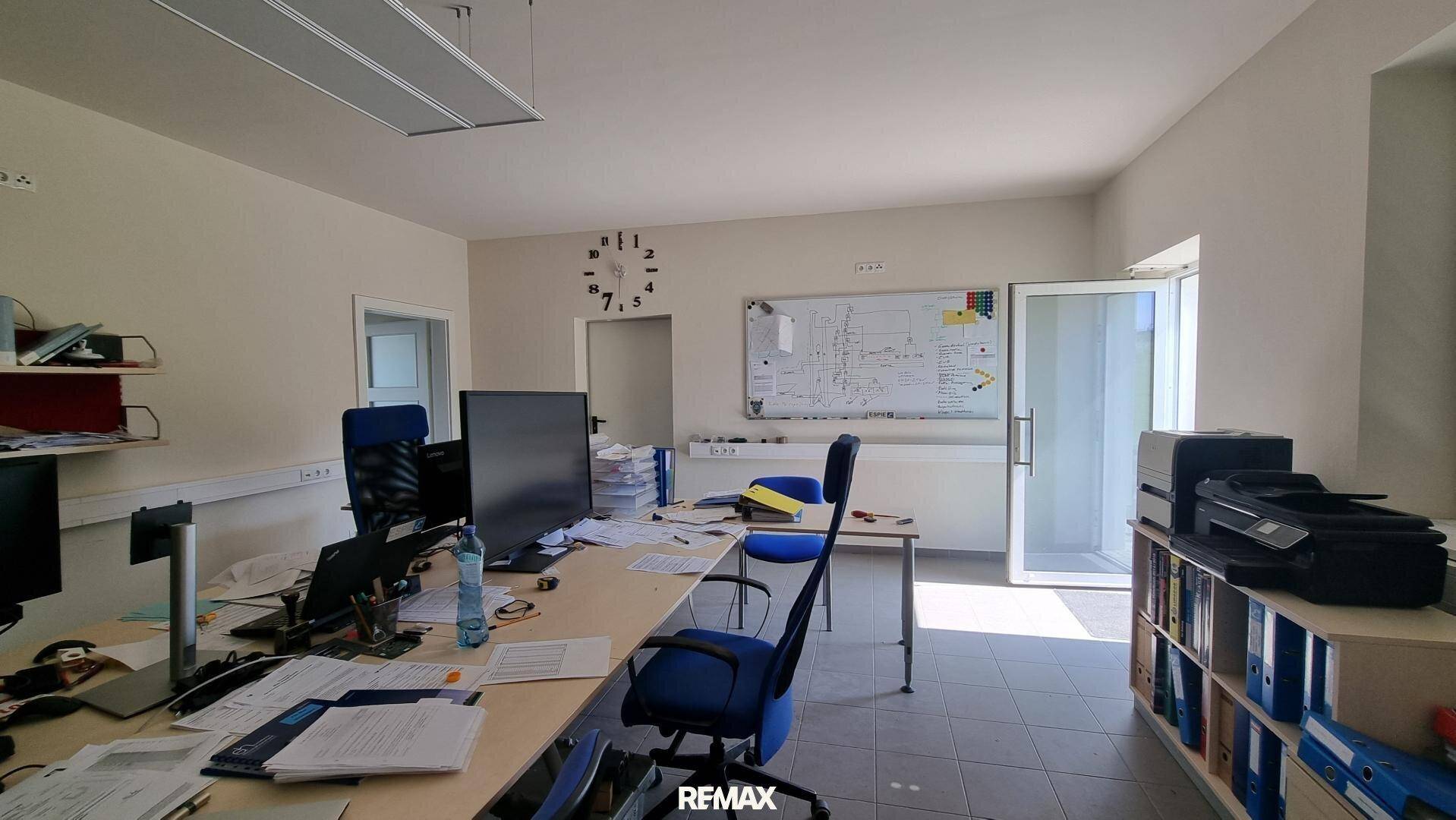 Büro mit Schreibtischen, Whiteboard und Fenstern, gut beleuchtet und funktional eingerichtet.