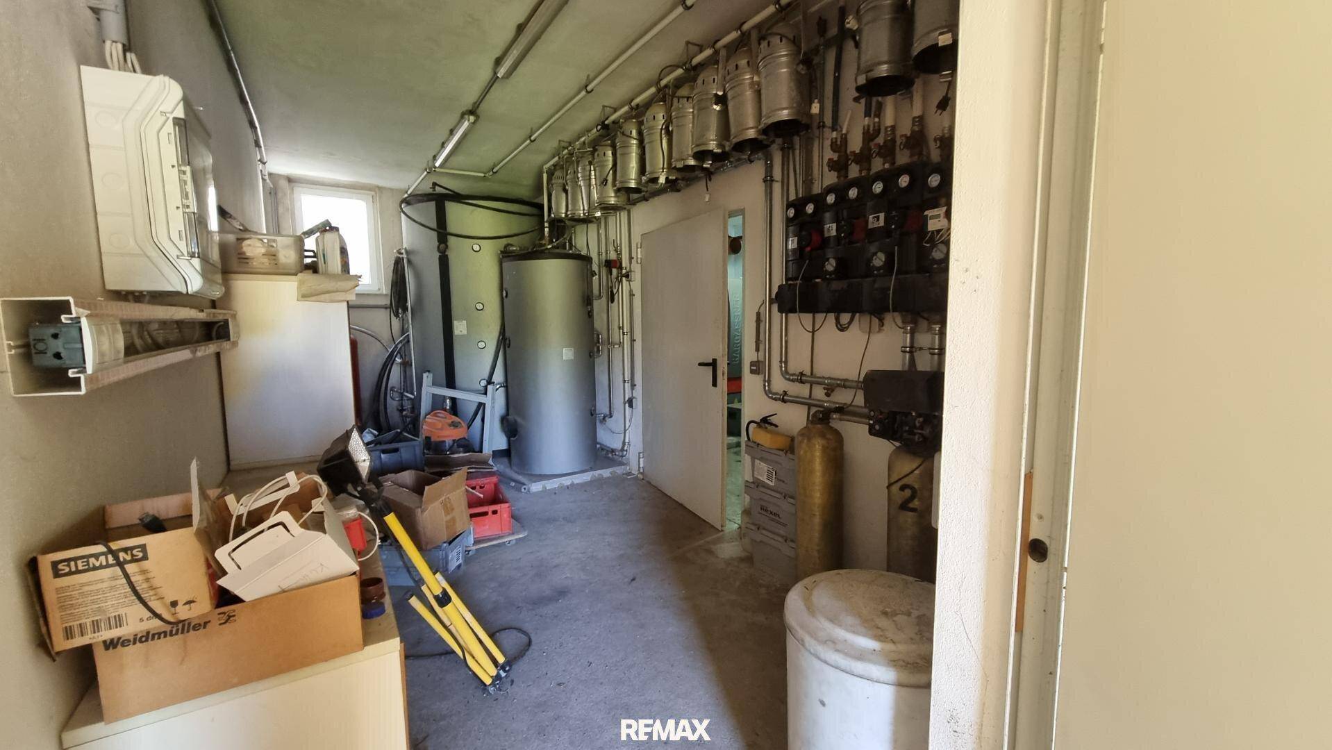 Technikraum mit Warmwasserspeichern, Rohren und verschiedenen Installationen, gut beleuchtet durch ein Fenster.
