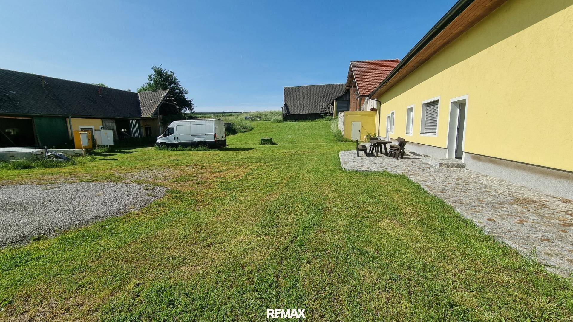 Weitläufiger Garten mit Rasenfläche, Gebäuden im Hintergrund und sonnigem Wetter, ländliche Umgebung.