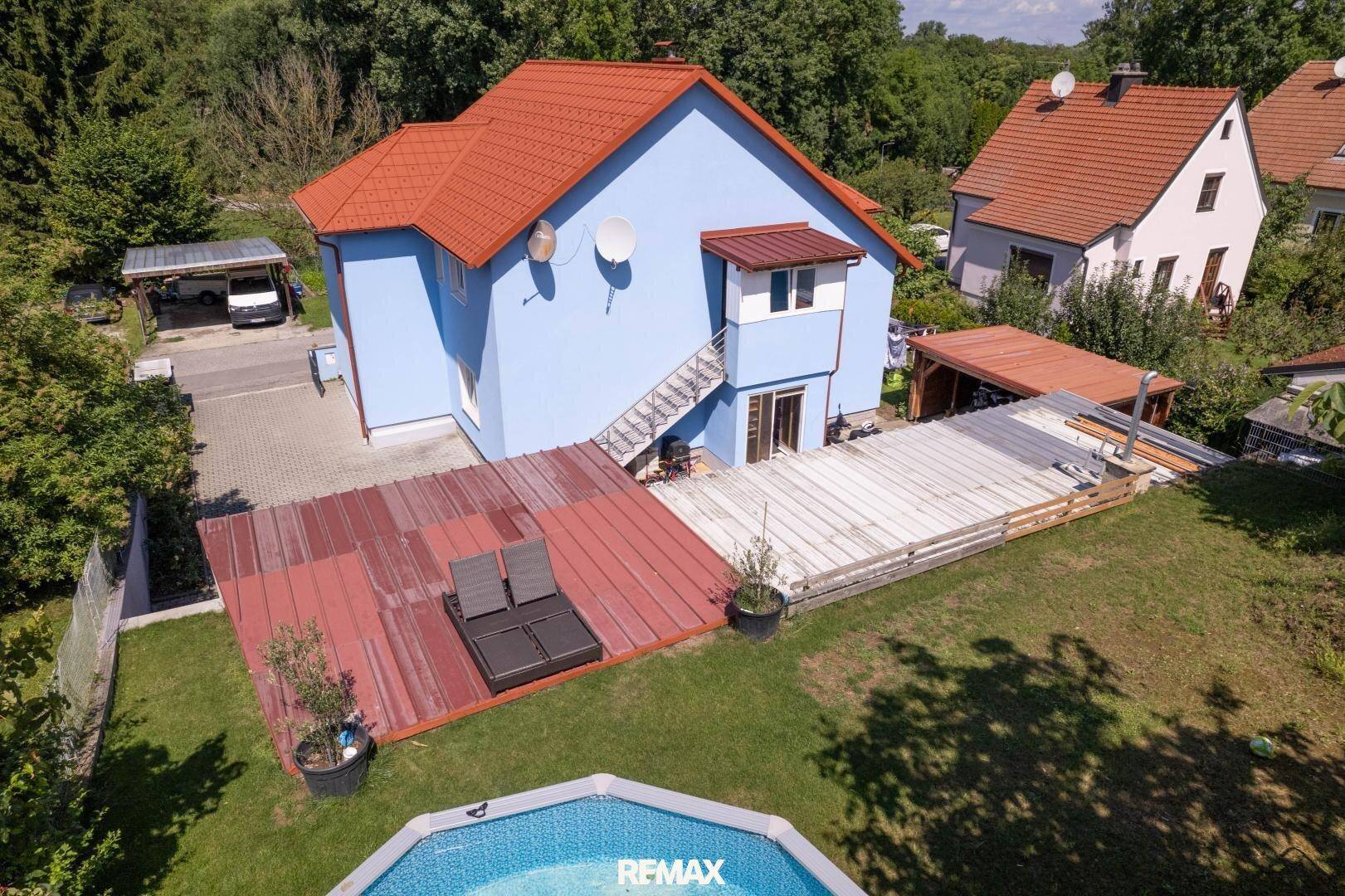 Blaues Einfamilienhaus mit rotem Dach, großer Terrasse, Garten und einem Aufstellpool.