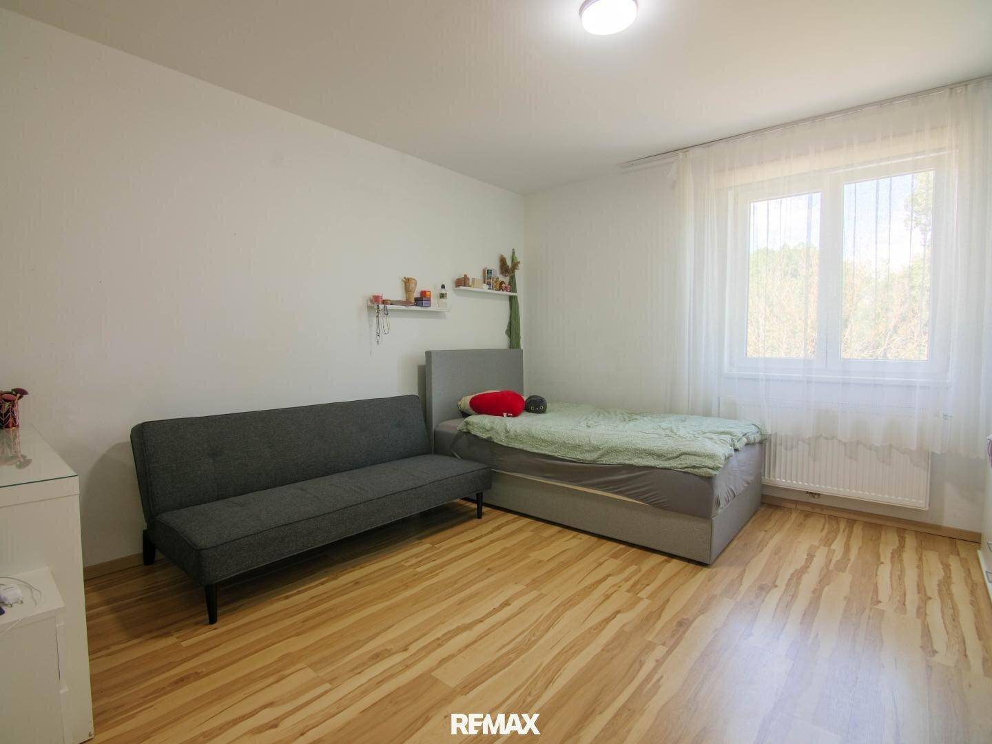 Schlafzimmer mit Einzelbett, Sofa und hellem Laminatboden, gut beleuchtet.