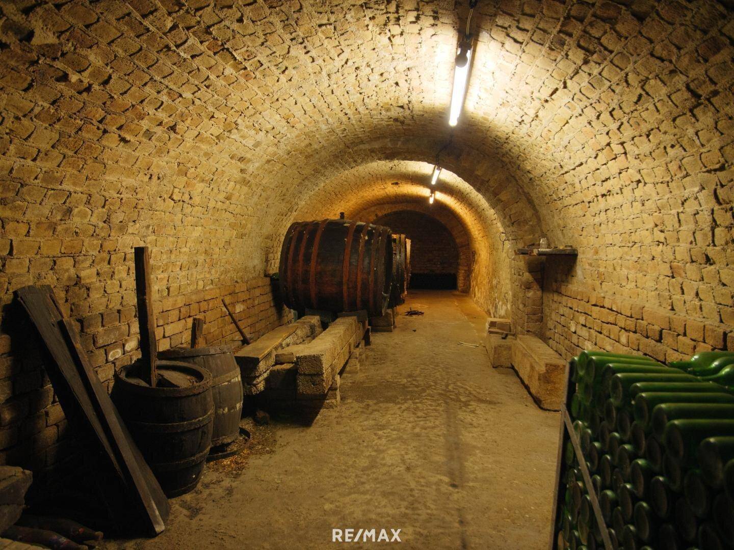 Langer Gewölbekeller mit Ziegelwänden, Weinfässern und Flaschenregalen.