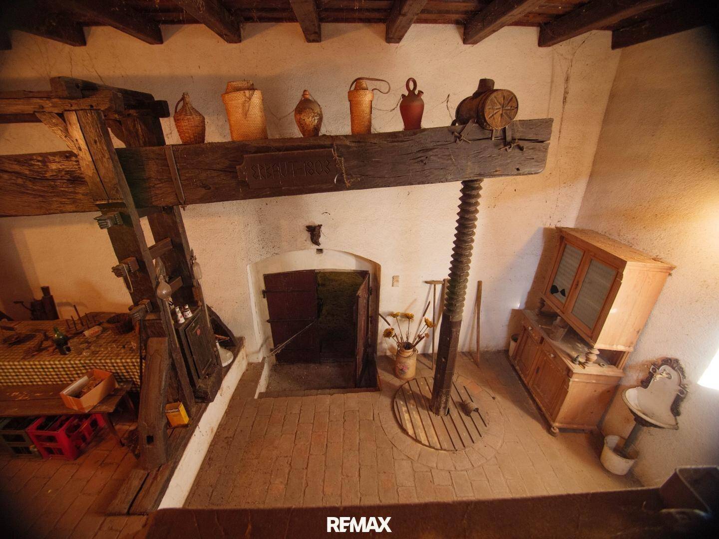 Historischer Weinkeller mit massiver Holzpresse, antiken Gefäßen und Ziegelboden.
