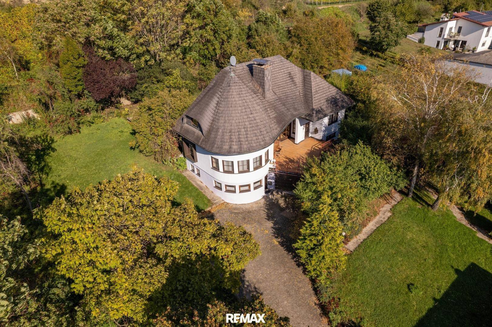 Luftaufnahme des Hauses mit markantem Dach, umgeben von Bäumen und einem gepflegten Garten.