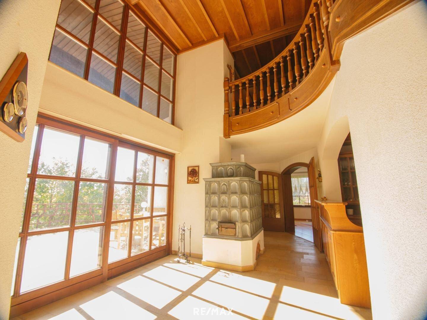 Heller Eingangsbereich mit hohen Fenstern, Holzdecke, gefliestem Boden und Kachelofen.