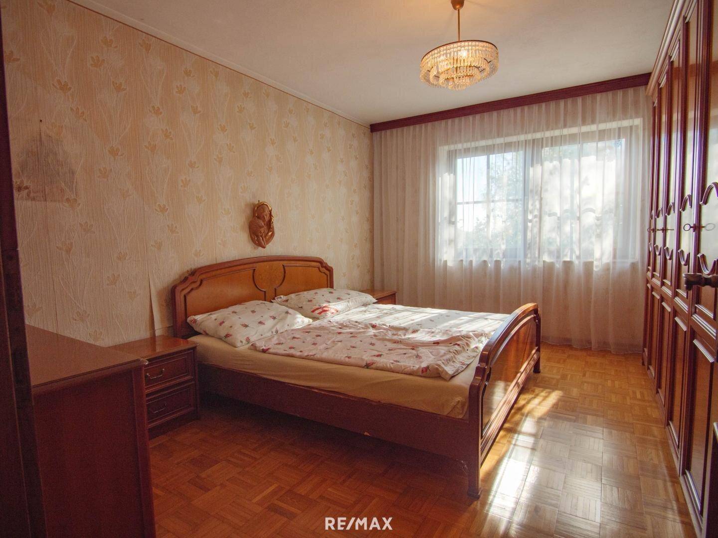 Schlafzimmer mit Doppelbett, Holzschrank, Parkettboden und gemusterter Tapete.