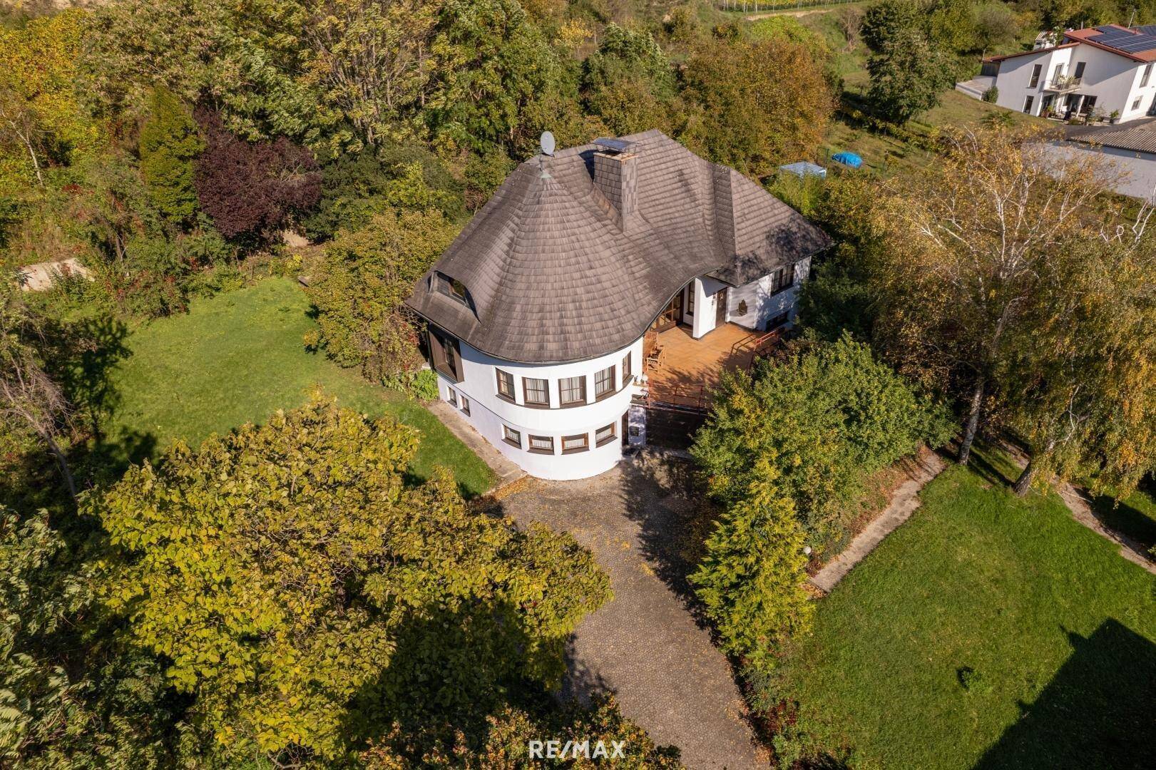 Luftaufnahme des Hauses mit geschwungenem Dach, umgeben von Bäumen und Grünflächen.
