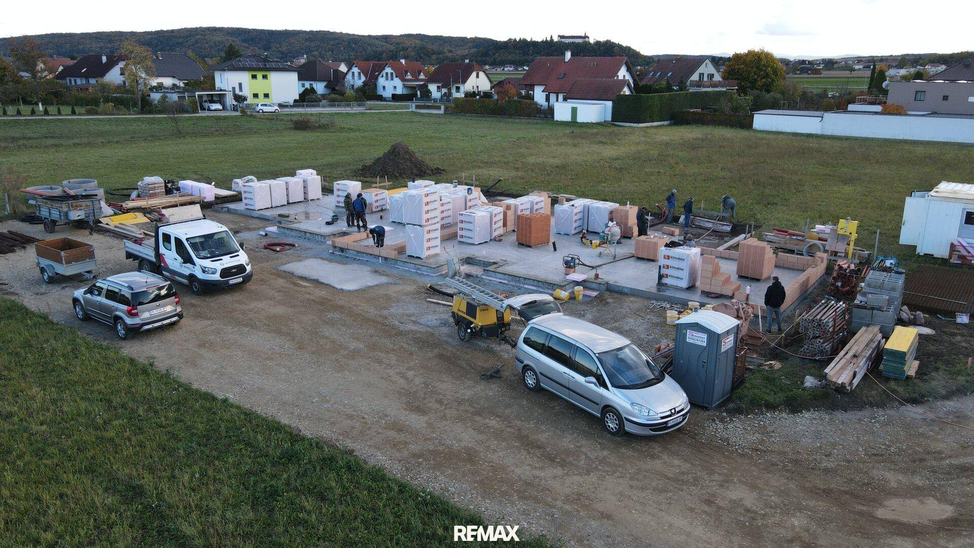 Luftaufnahme einer Baustelle mit beginnendem Mauerwerk und Materiallieferungen.