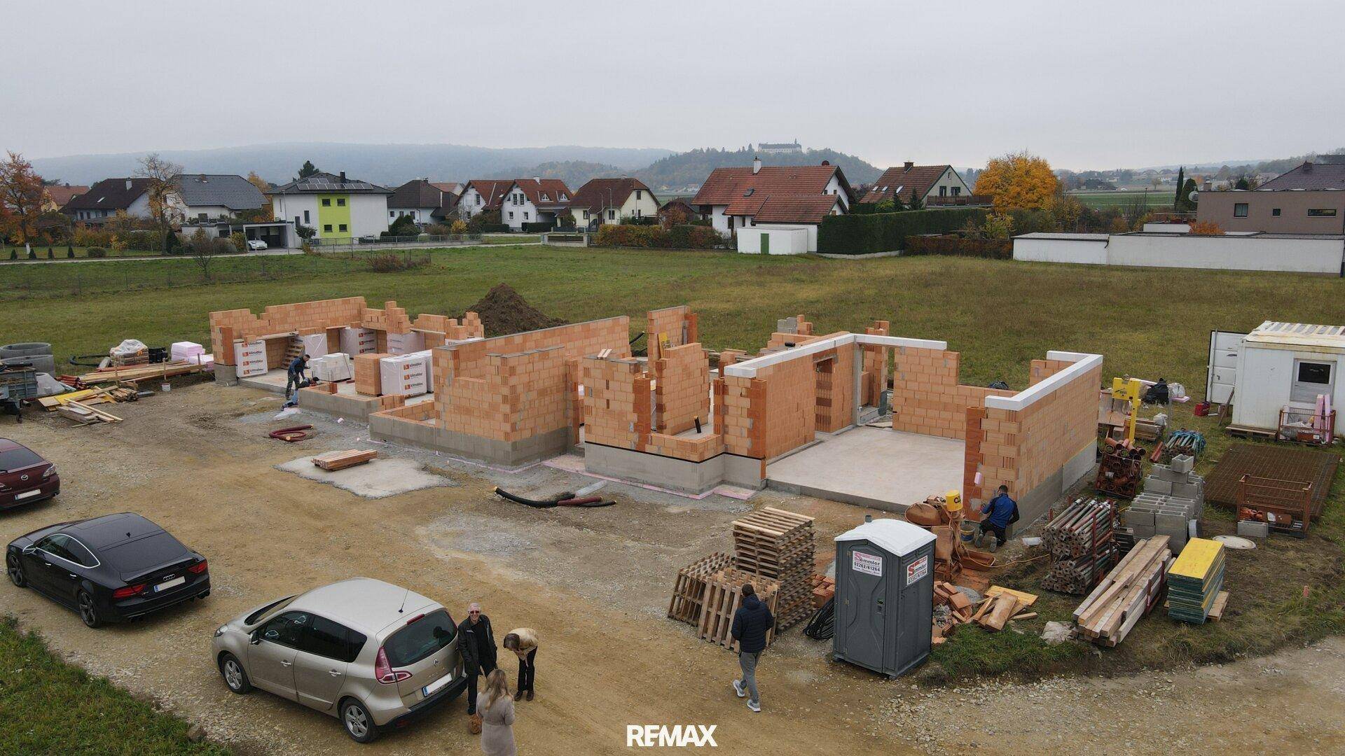 Luftaufnahme einer Baustelle mit Rohbau und Arbeitern, umgeben von ländlicher Landschaft.