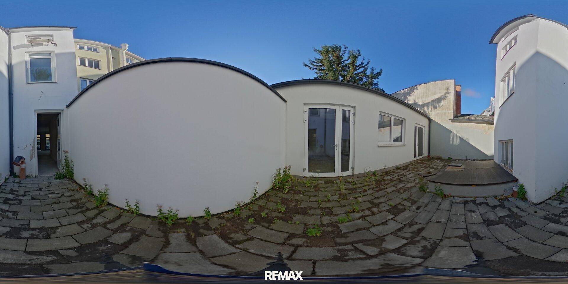 Panoramaansicht eines gepflasterten Innenhofs zwischen weißen Gebäudeteilen mit blauem Himmel.