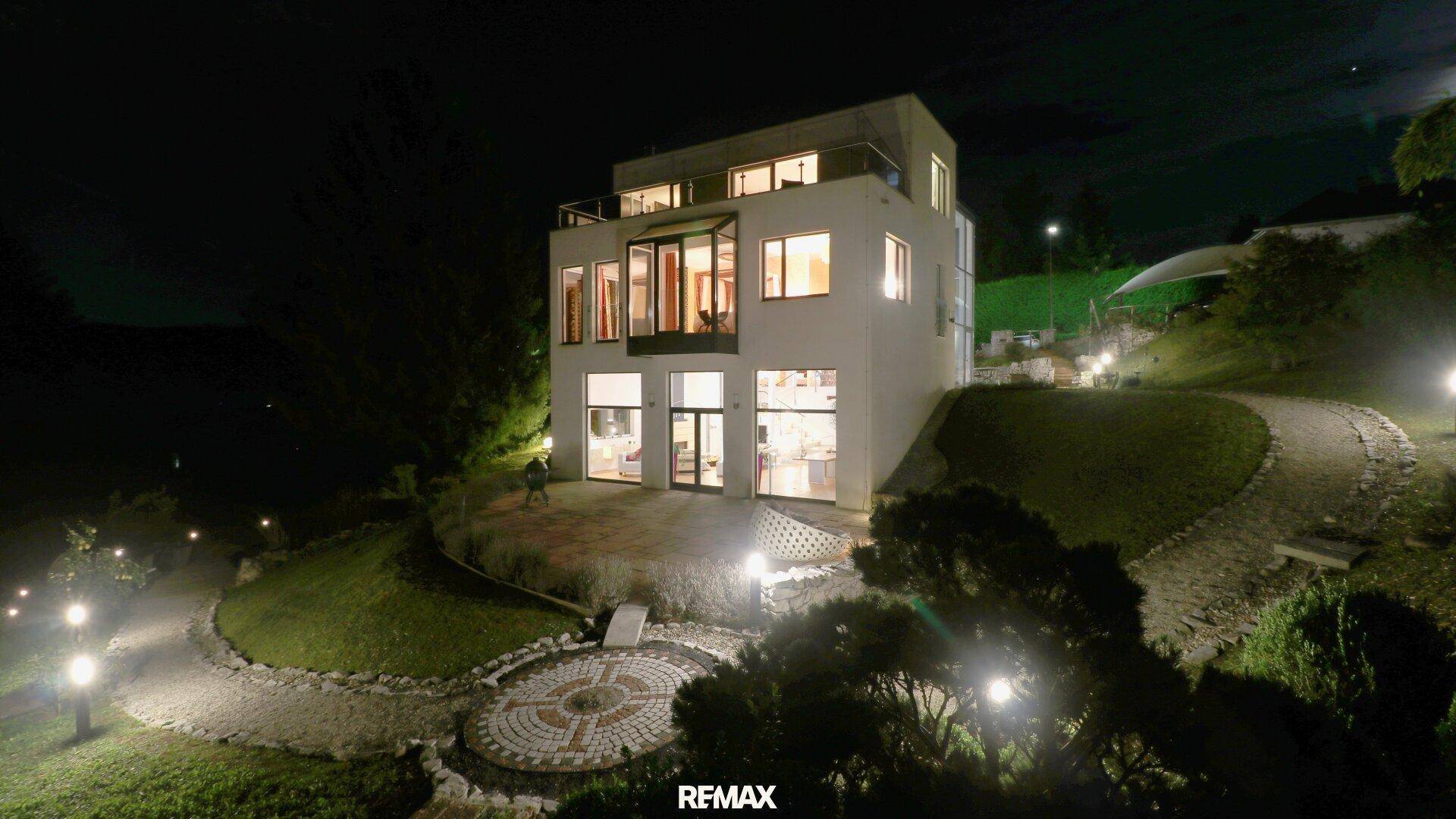 Modernes Gebäude bei Nacht, beleuchtet, mit stilvoll angelegtem Garten und Wegen.