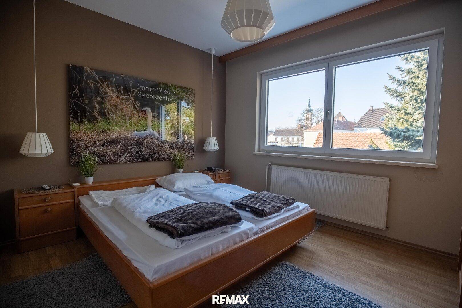 Schlafzimmer mit Doppelbett, Nachttischen und großem Fenster, das viel Tageslicht hereinlässt.