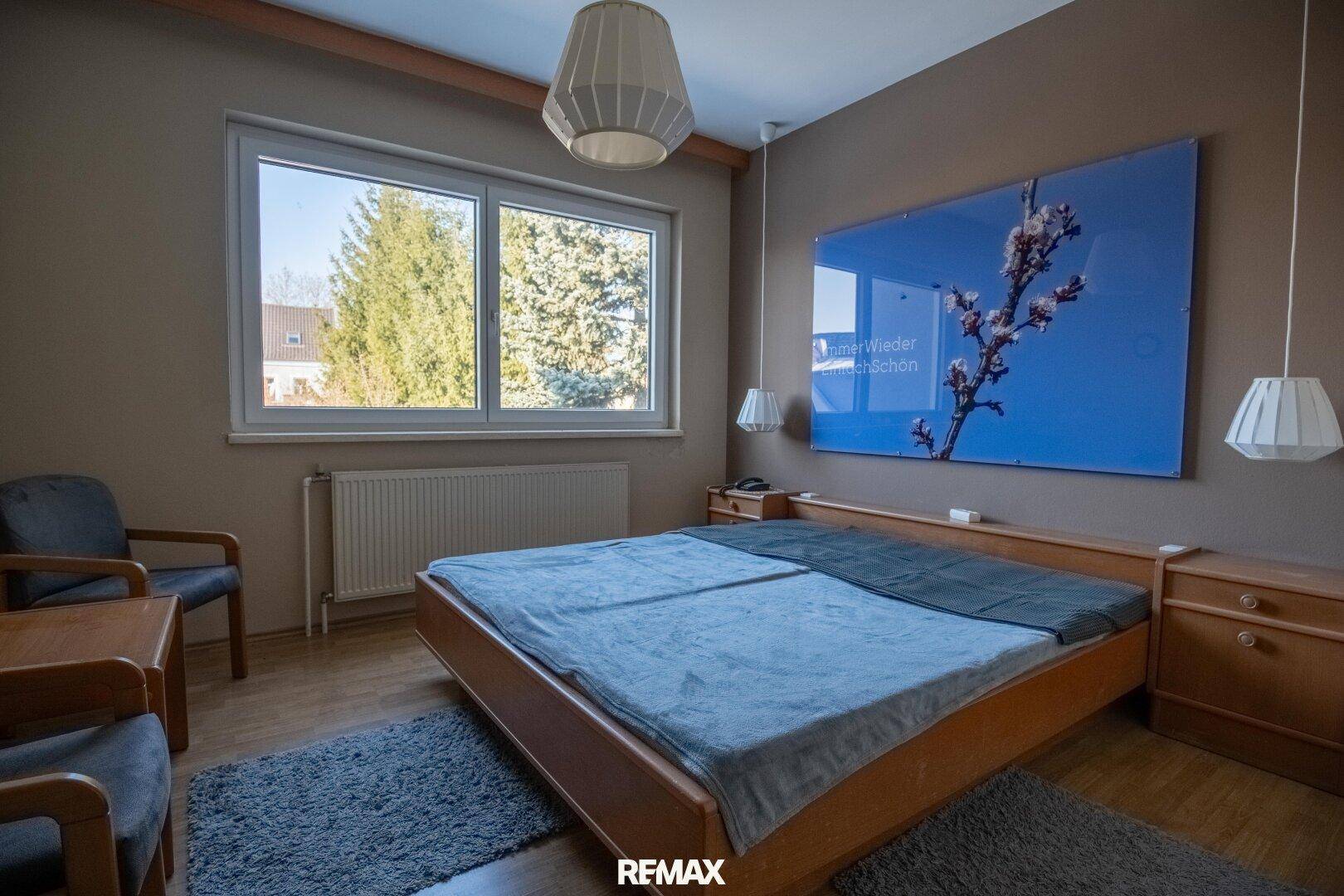 Schlafzimmer mit Doppelbett, Nachttischen und großem Fenster, das viel Tageslicht hereinlässt.