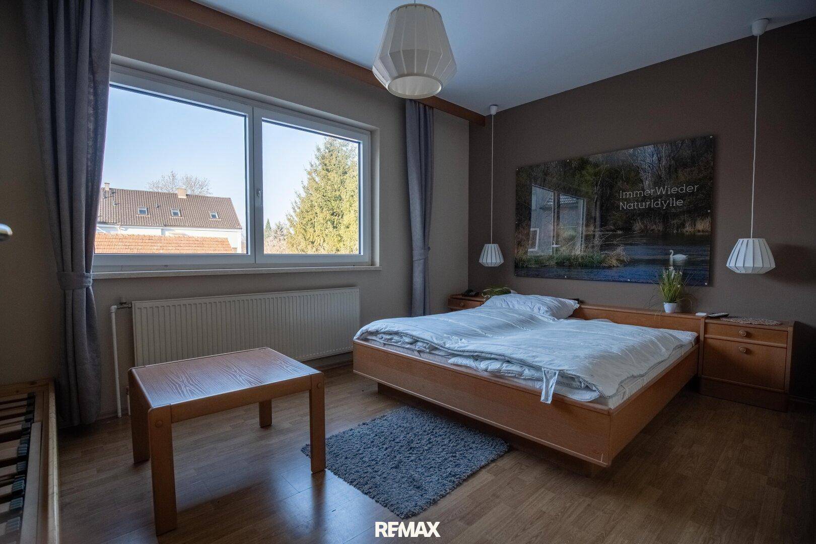 Schlafzimmer mit Doppelbett, Nachttischen und großem Fenster, das viel Tageslicht hereinlässt.