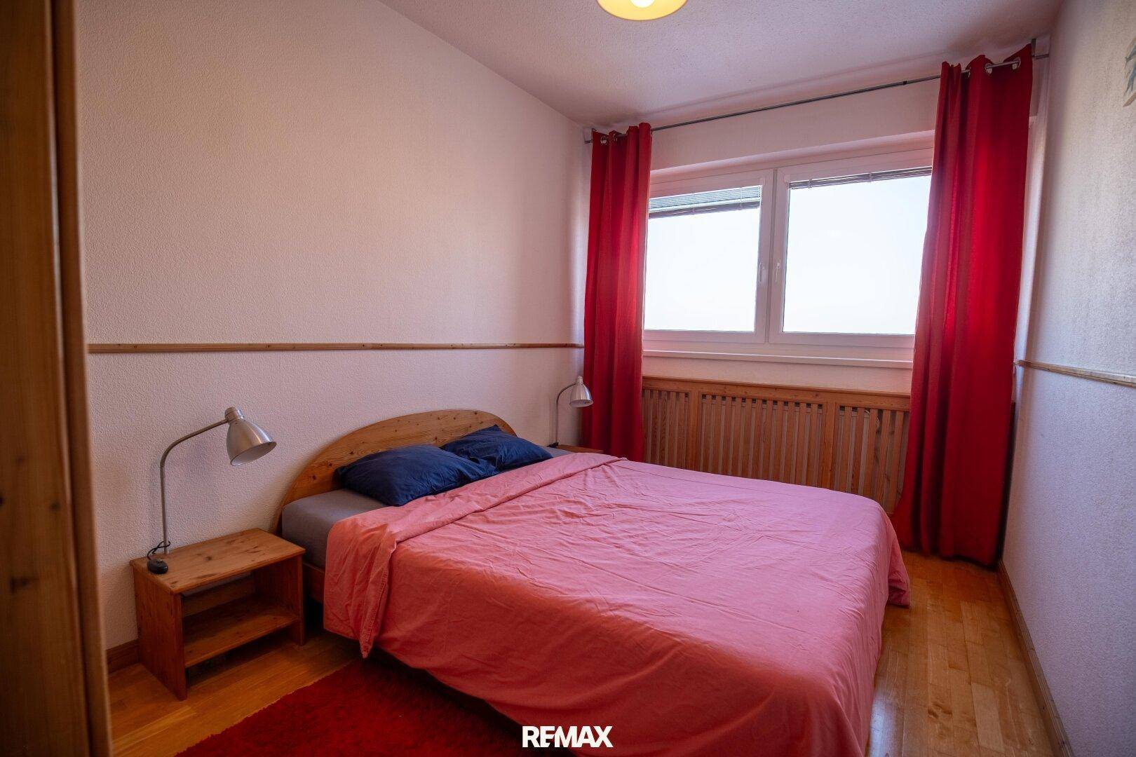 Schlichtes Schlafzimmer mit Holzboden, Doppelbett und roten Vorhängen am Fenster.
