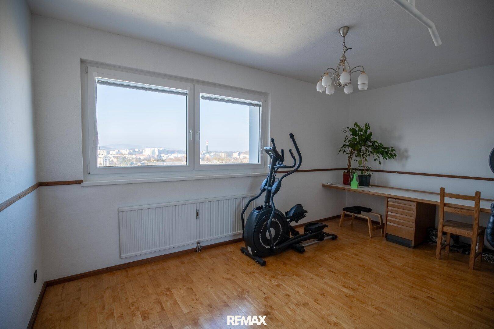 Heller Fitnessraum mit Trainingsgerät, Schreibtisch und weitem Blick über die Stadt.