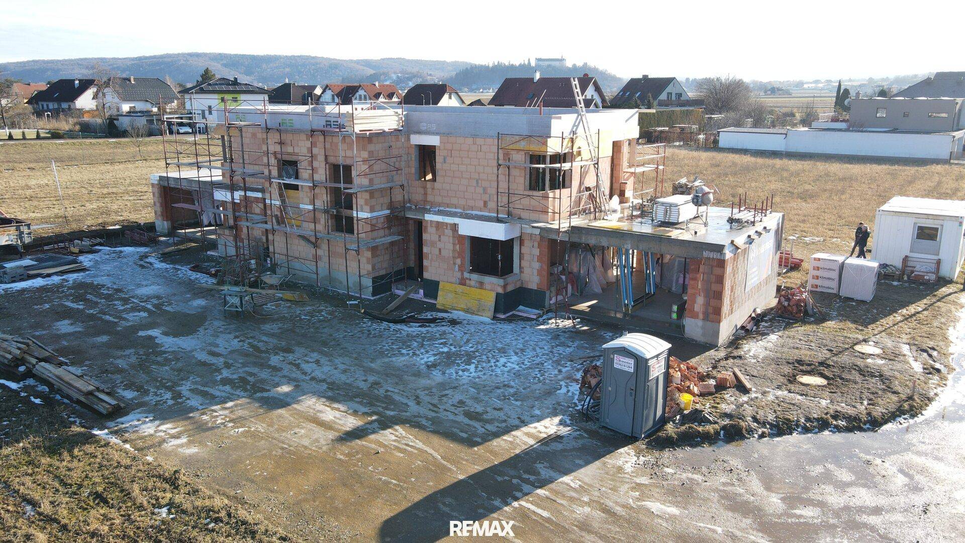 Luftaufnahme einer Baustelle mit Rohbau, Gerüst und Schneeresten auf dem Gelände.