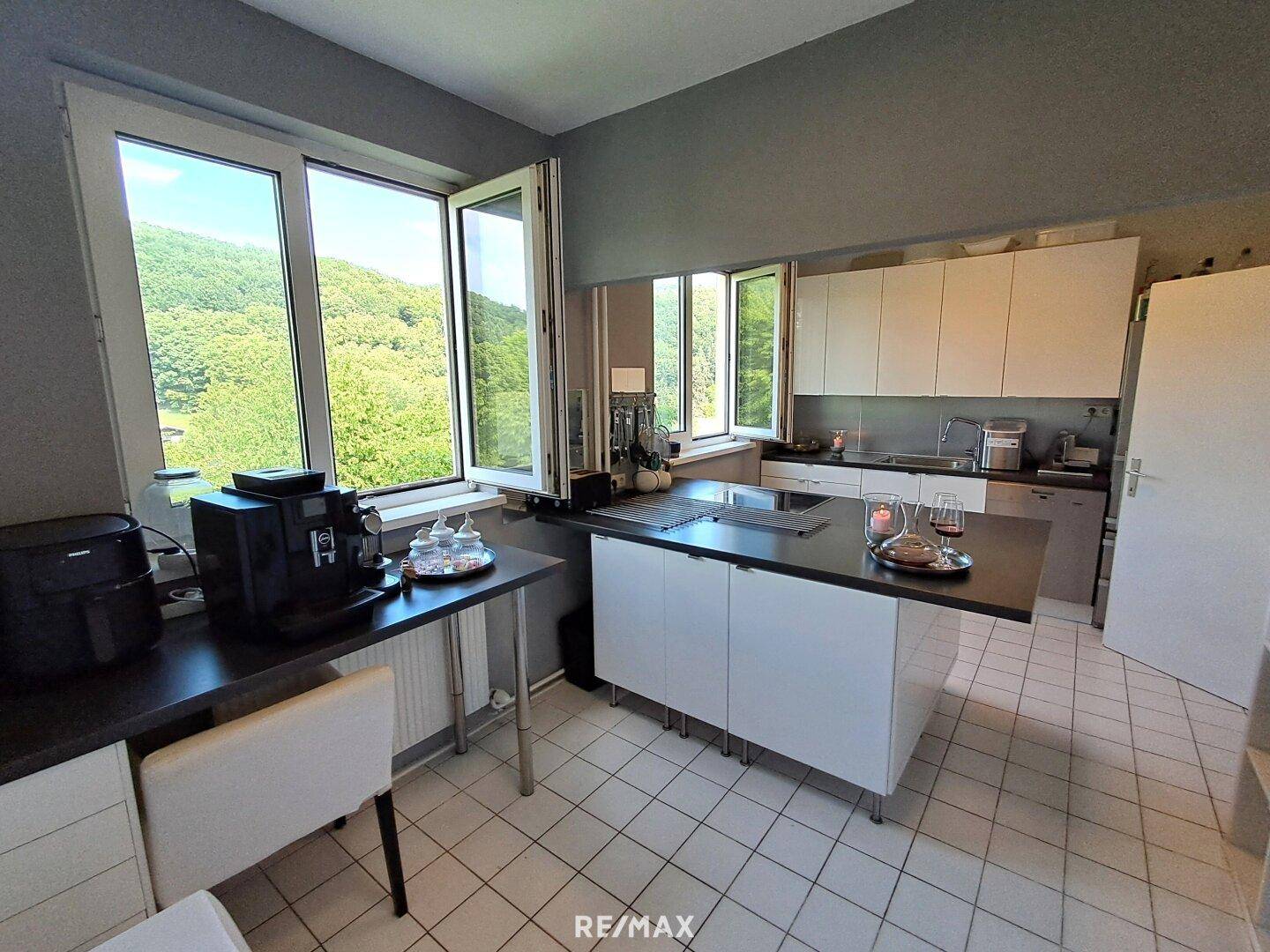 Moderne Küche mit dunkler Arbeitsplatte, weißen Schränken und großen Fenstern mit Waldblick.