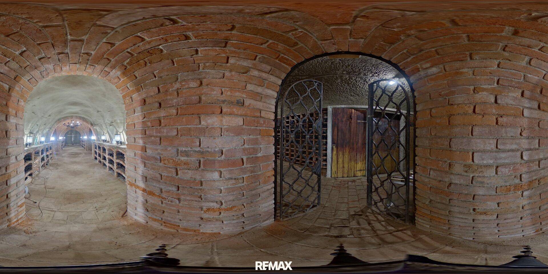 Panoramablick in einen Weinkeller mit Ziegelgewölben und einem dekorativen Metalltor am Eingang.