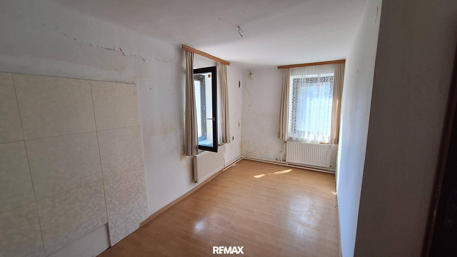 Unmöbliertes Zimmer mit Laminatboden, zwei Fenstern und sichtbaren Gebrauchsspuren.