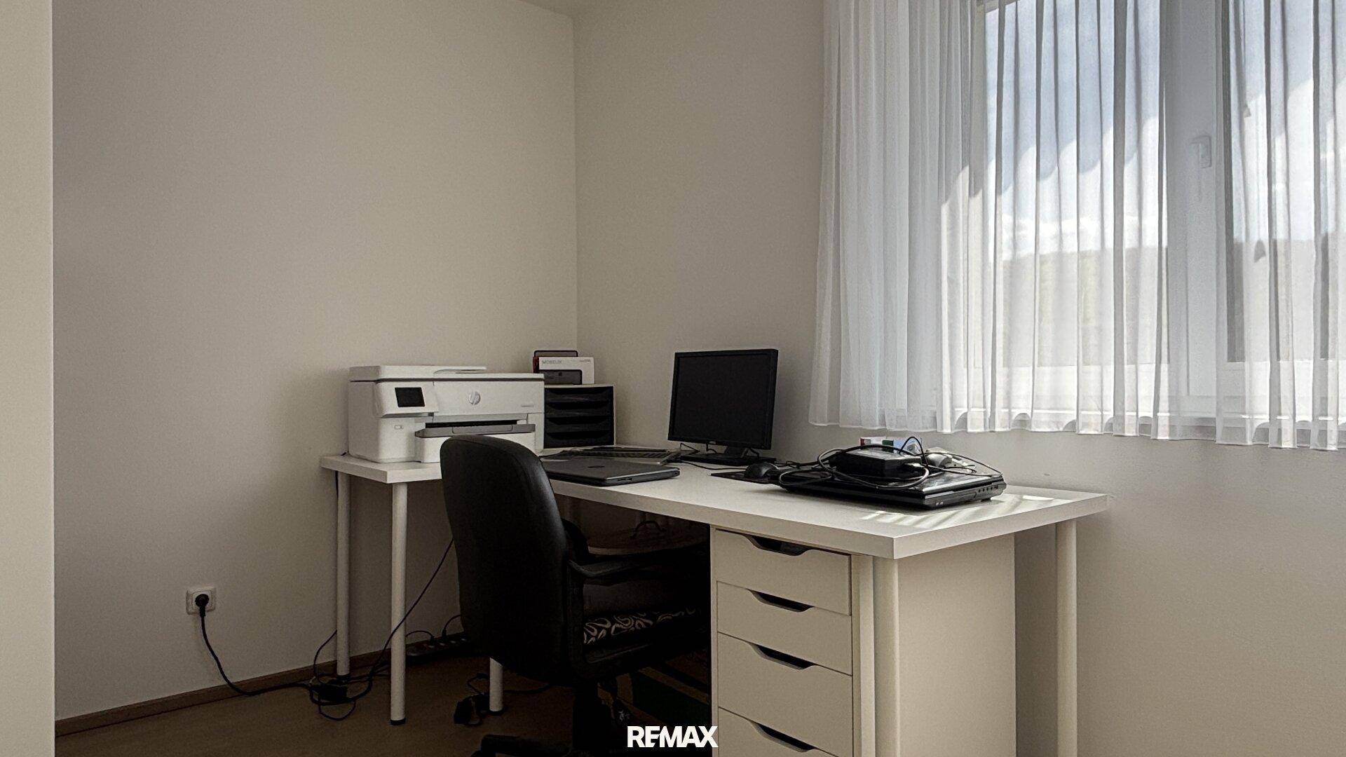 Funktionales Arbeitszimmer mit Schreibtisch, Computer und Drucker, hell durch großes Fenster.